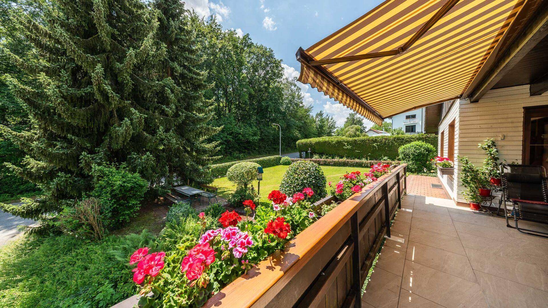 Blick von der Terrasse auf den gepflegten Garten mit Blumenkästen und hohen Bäumen.