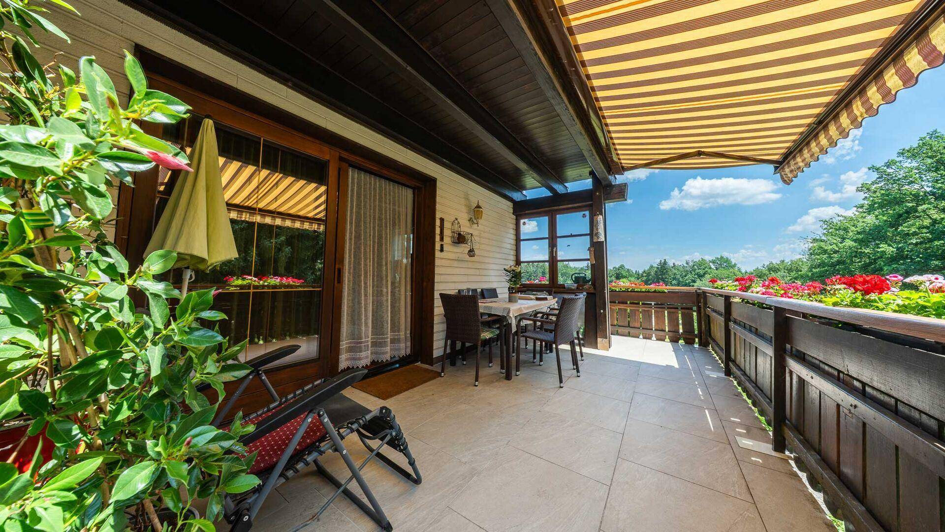 Geräumige Terrasse mit Esstisch, Sonnenliege und ausziehbarer Markise, umgeben von Pflanzen.