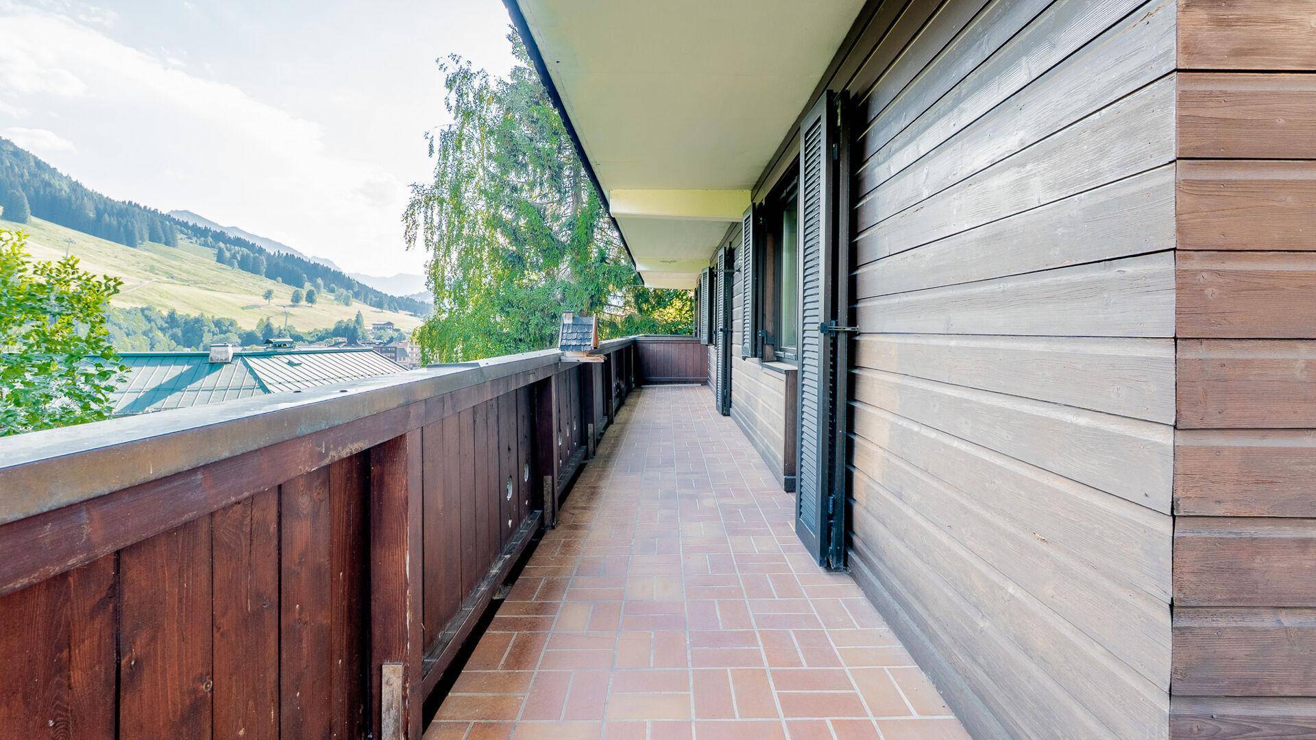 Langer Balkon mit Holzgeländer und Terrakottafliesen, umgeben von grüner Natur.