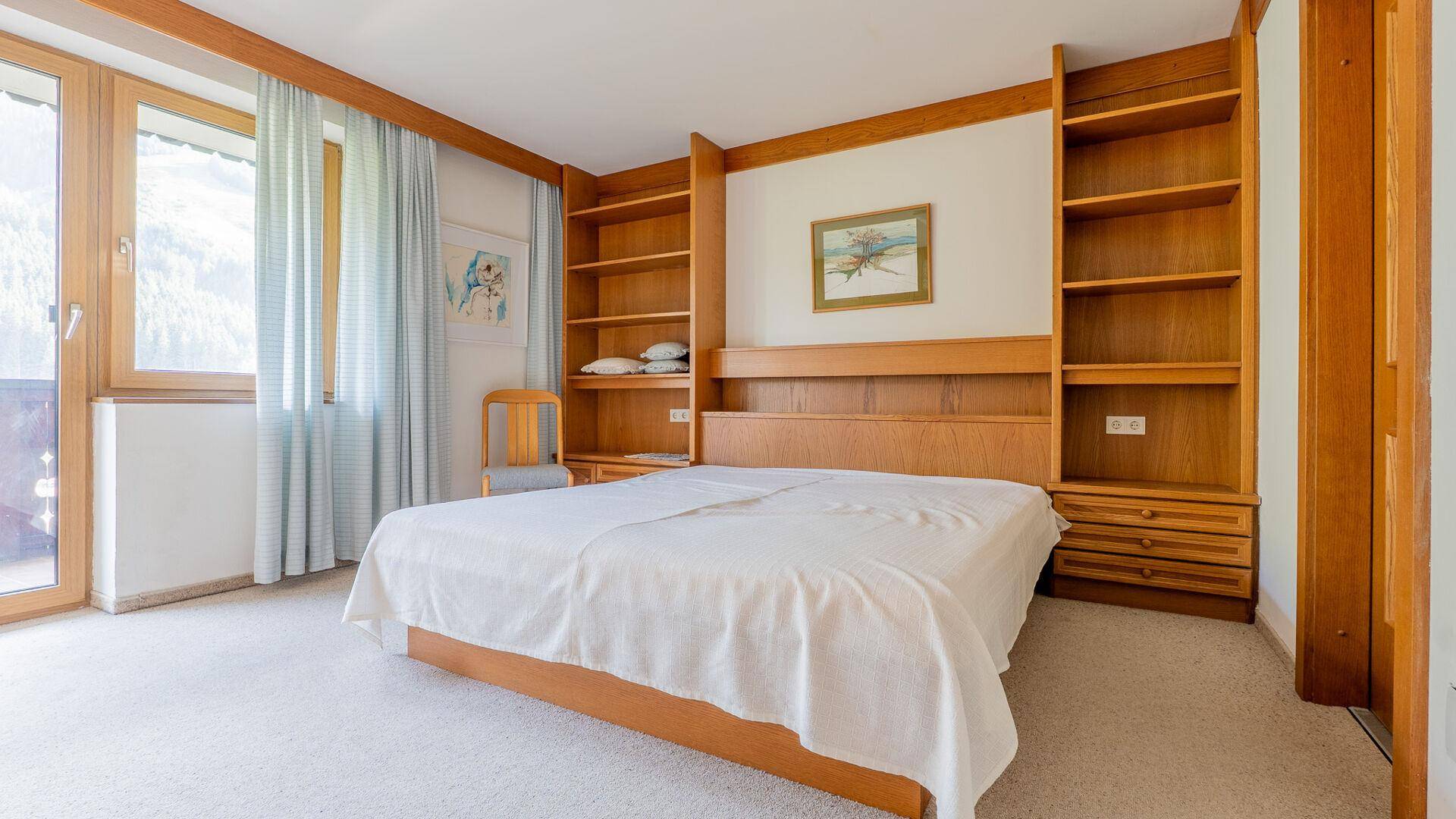 Schlafzimmer mit Doppelbett, Teppichboden, eingebauten Holzregalen und Zugang zu einem Balkon.