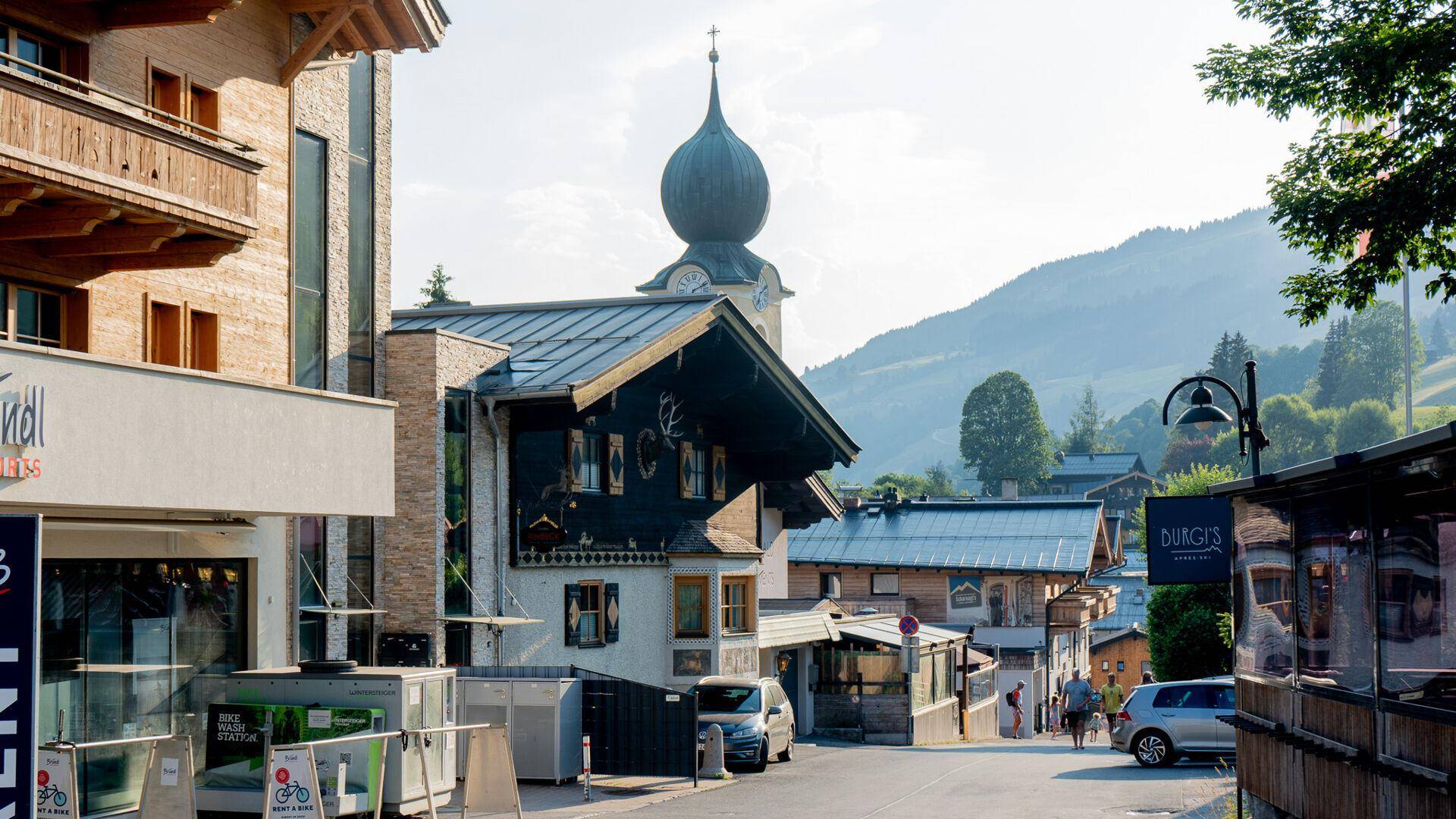 Belebte Dorfstraße mit traditionellen Gebäuden, einem Kirchturm und Bergen im Hintergrund.
