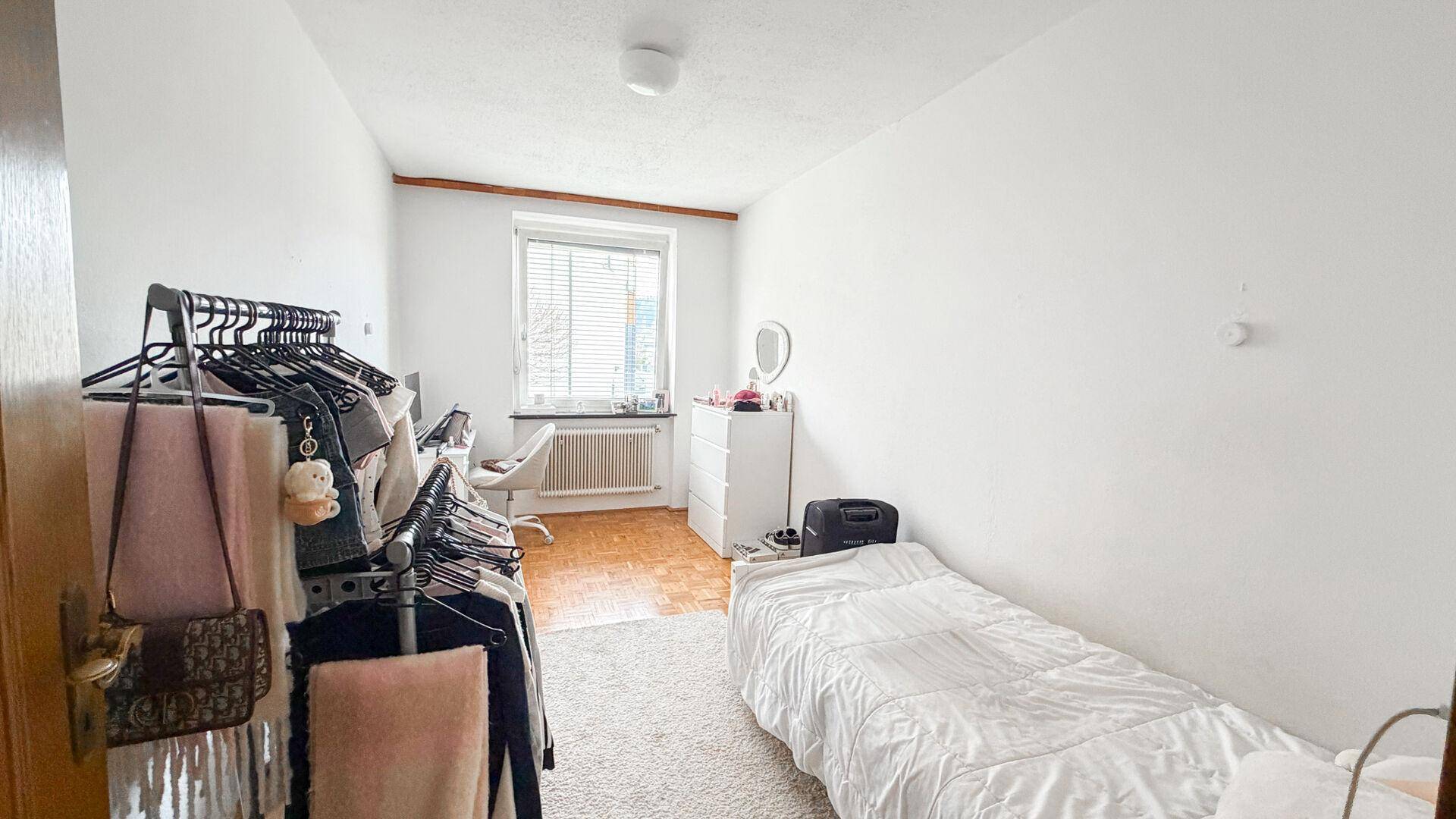 Kleines Schlafzimmer oder Gästezimmer mit Einzelbett, Kleiderstange und Fenster, einfach eingerichtet.