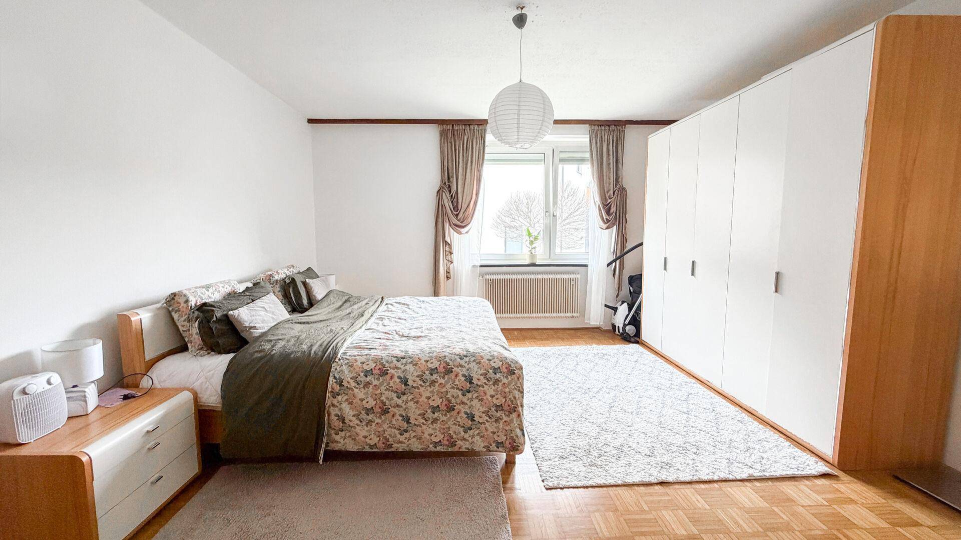 Helles Schlafzimmer mit Doppelbett, großem Kleiderschrank und Parkettboden, gut beleuchtet.