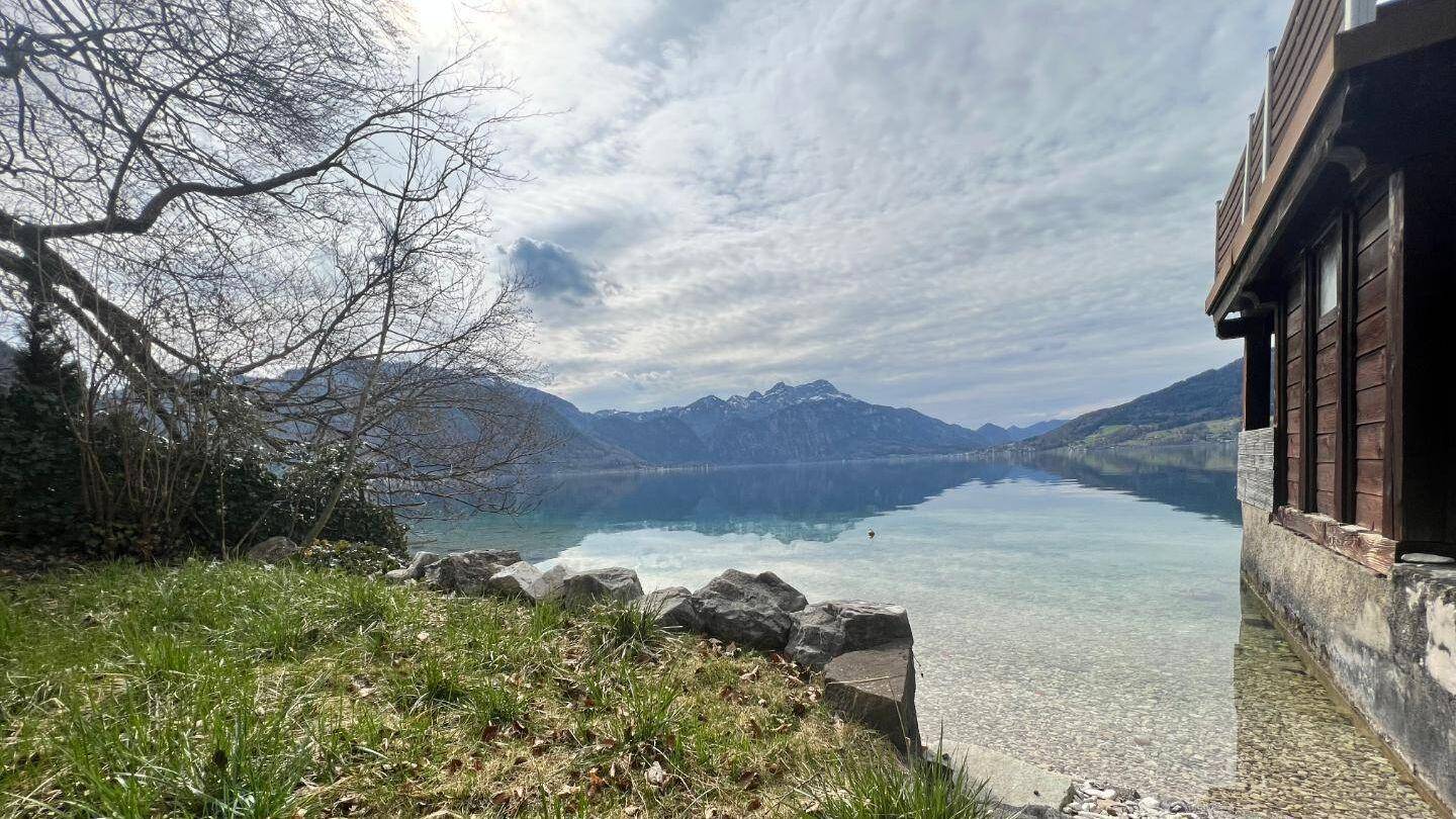 Ruhiger Seeuferbereich mit klarem Wasser, umgeben von Bergen und einem Holzgebäude am Rand.