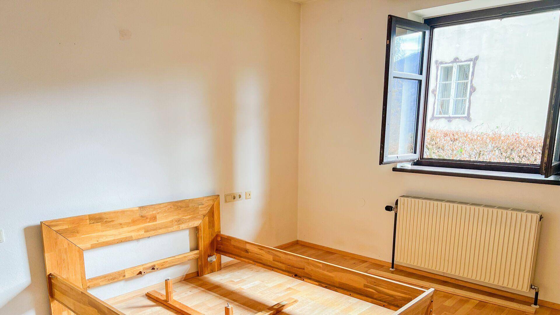 Schlichtes Schlafzimmer mit Holzbettrahmen, weißen Wänden und Fenster mit Heizkörper.