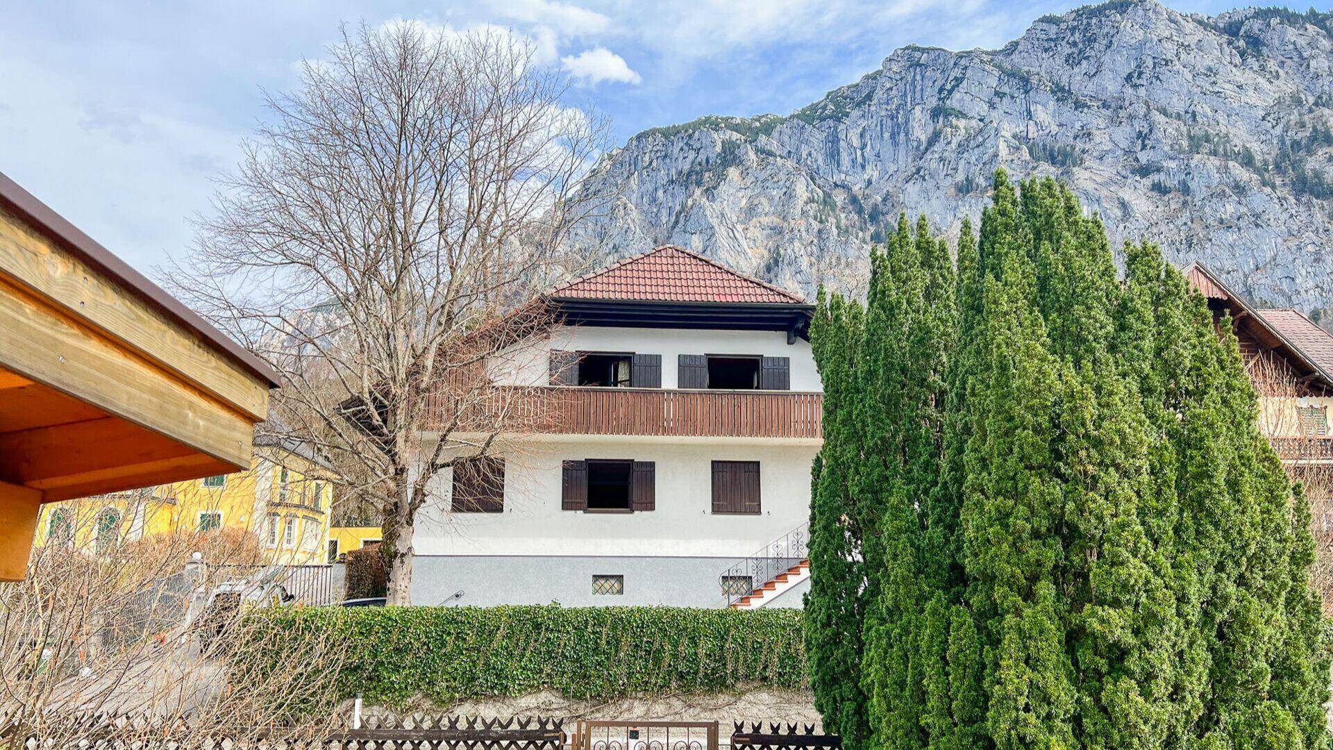 Traditionelles Haus mit rotem Ziegeldach und braunen Fensterläden vor einer imposanten Bergkulisse.