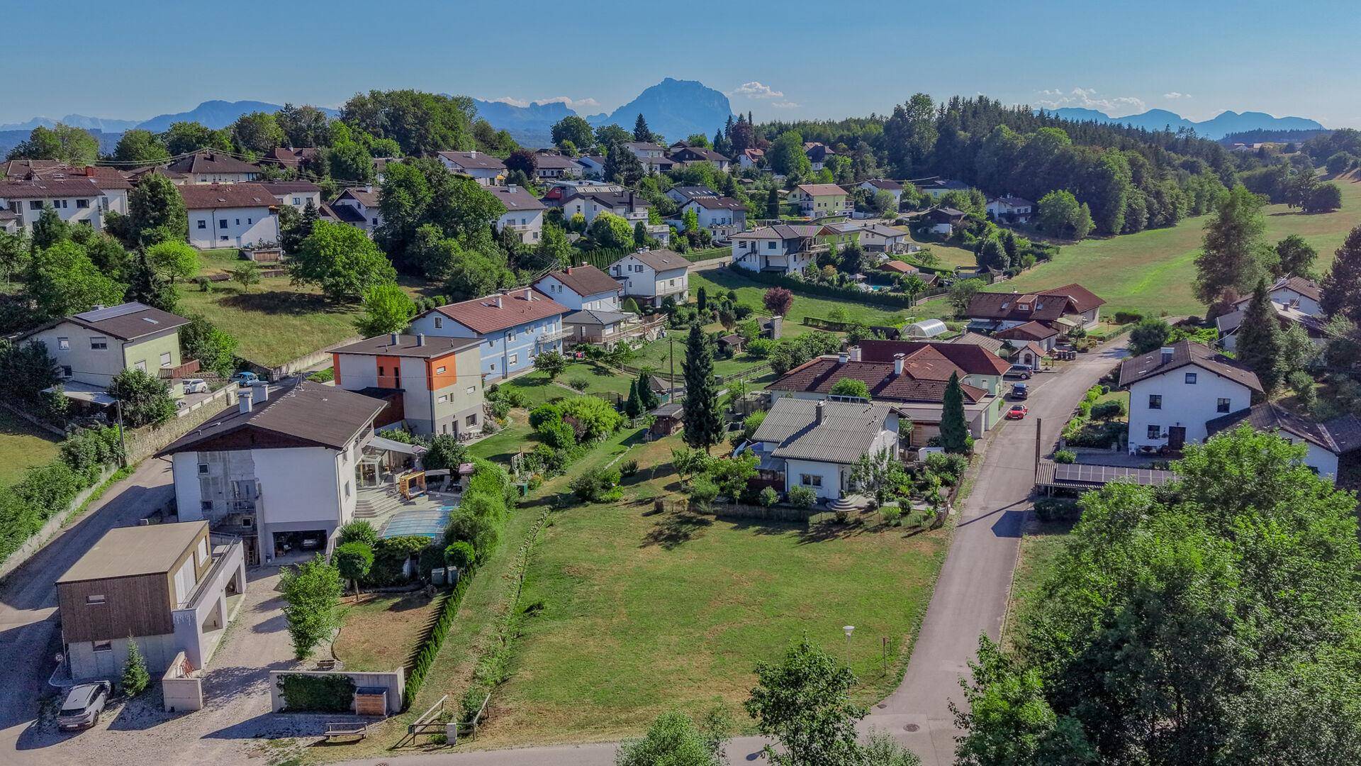 Weitläufige Luftaufnahme einer ländlichen Siedlung eingebettet in eine grüne Hügellandschaft mit Bergen im Hintergrund.