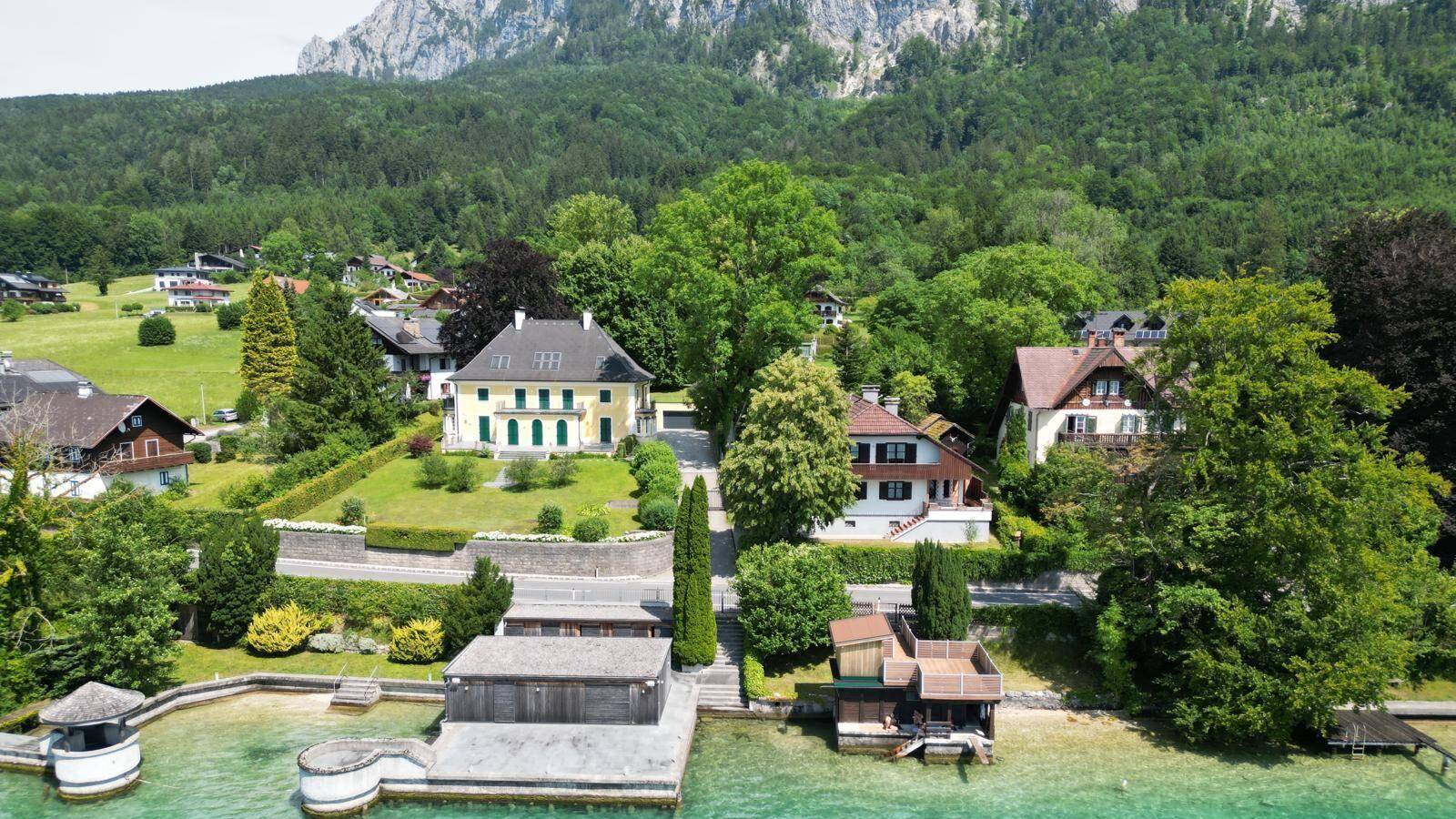 Luftaufnahme von mehreren Häusern am Seeufer, eingebettet in grüne Natur und Berge.