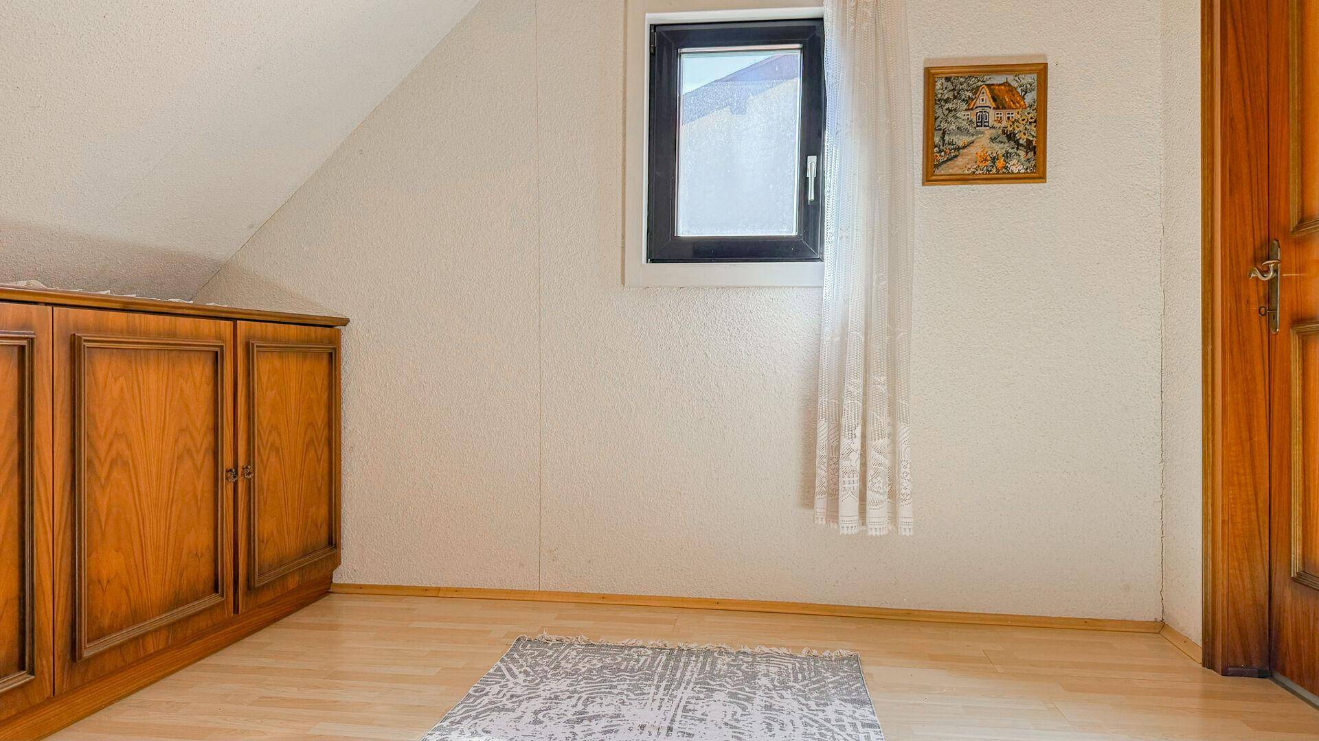 Dachzimmer mit schräger Decke, Fenster und traditionellem Holzschrank.