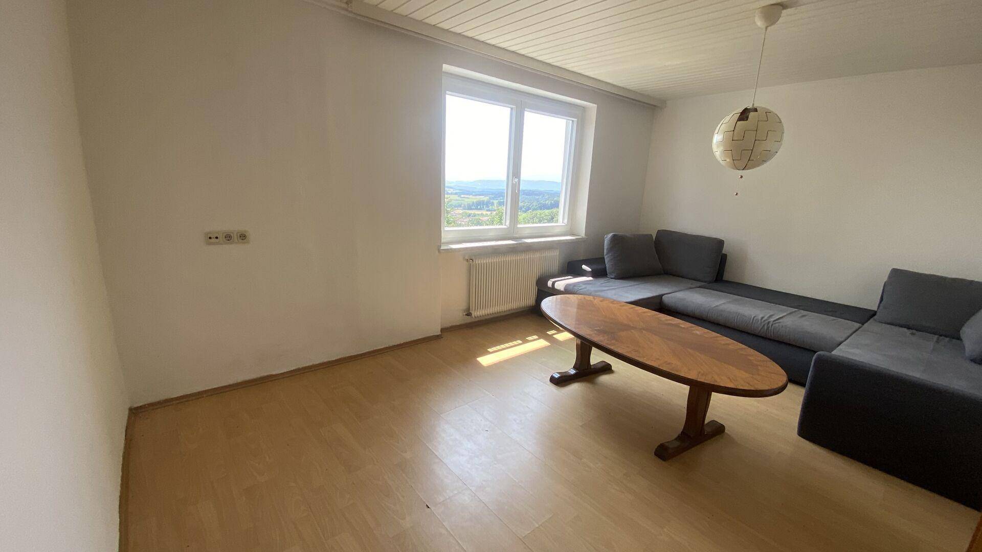 Geräumiges Wohnzimmer mit hellem Laminatboden, Sofa und einem Fenster mit Blick ins Grüne.