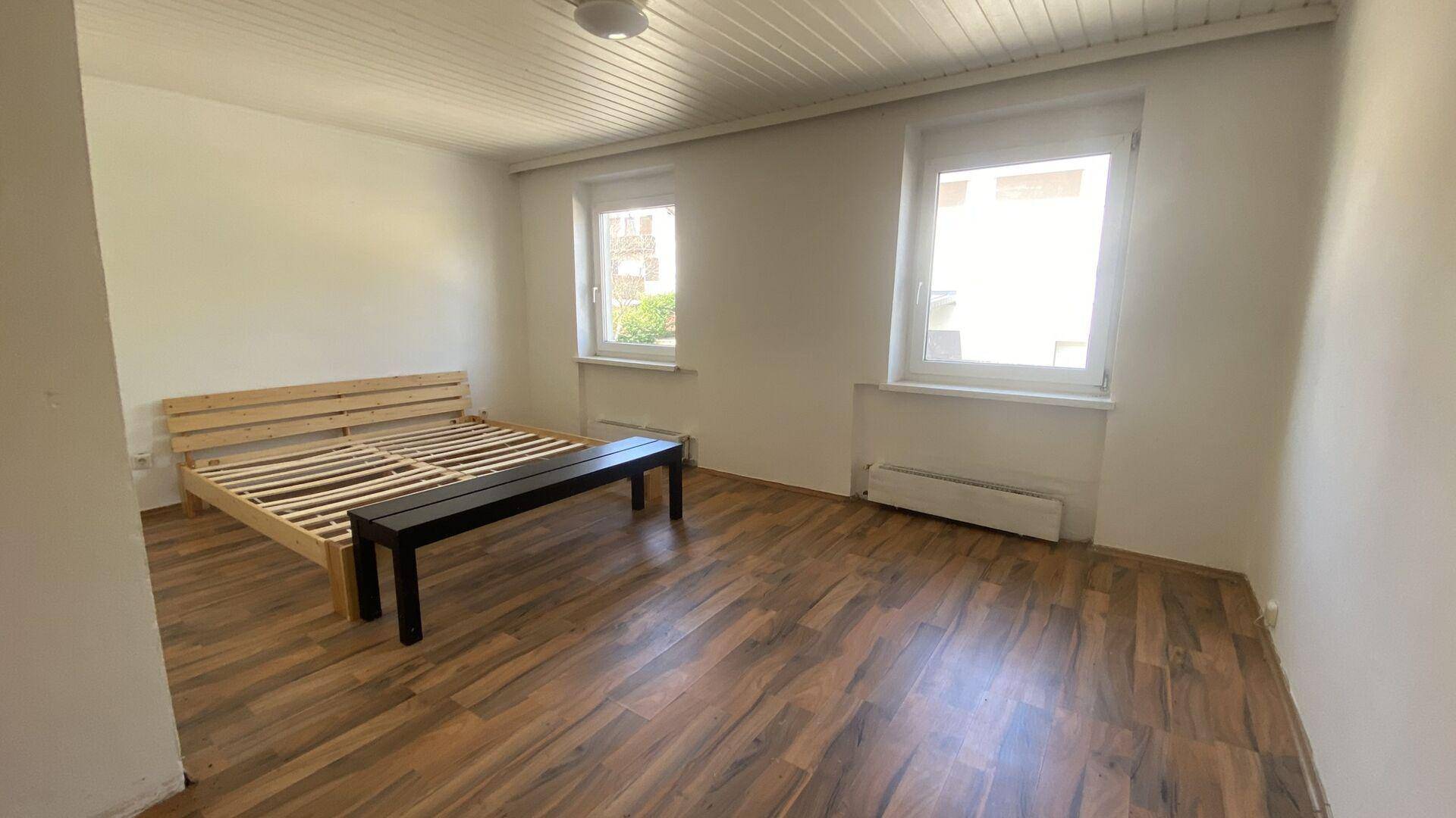 Schlafzimmer mit einfachem Holzbettrahmen und dunklem Laminatboden, hell durch zwei Fenster.