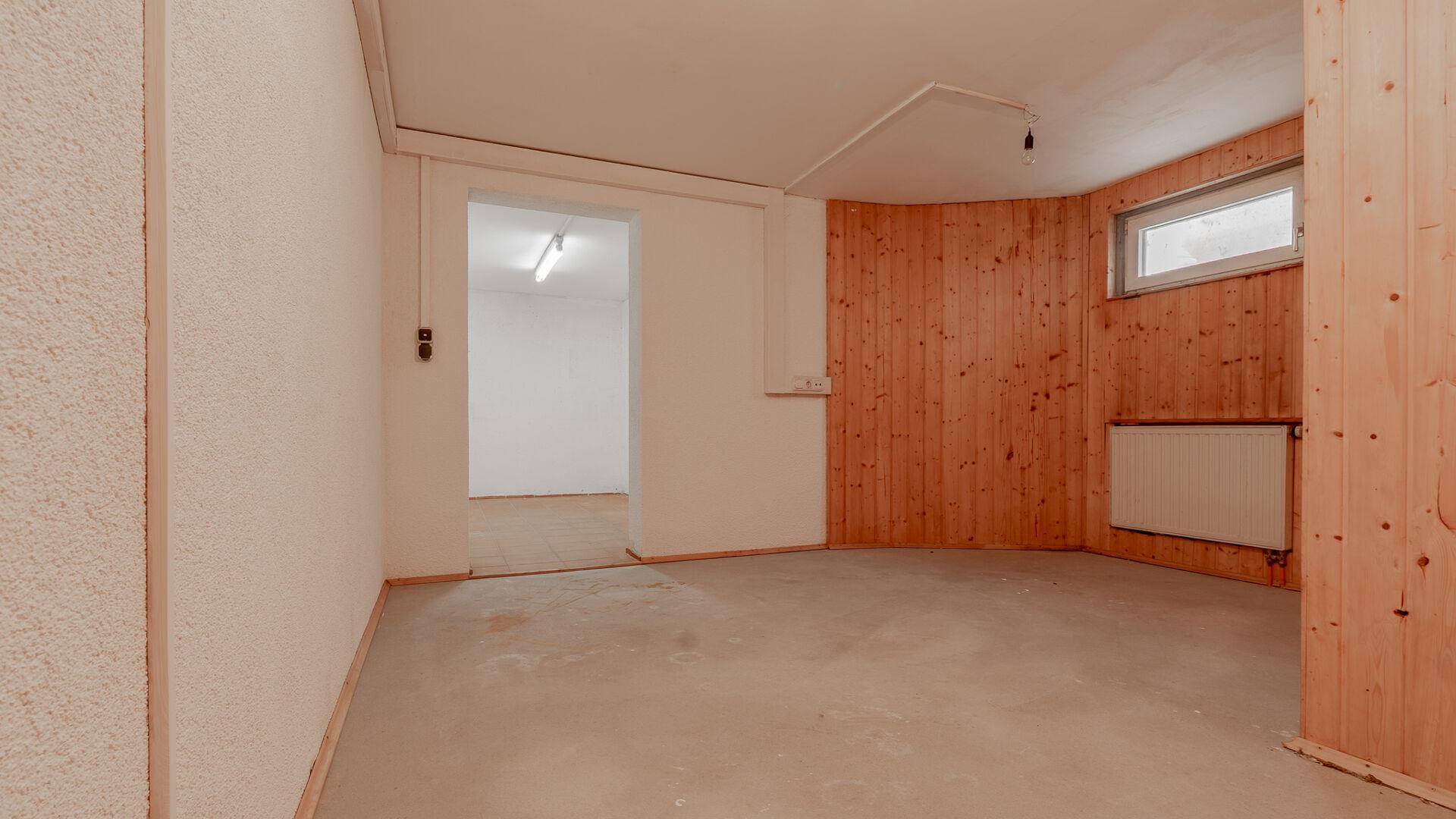 Kellerraum mit Betonboden, Holzverkleidung an einer Wand und kleinem Fenster.