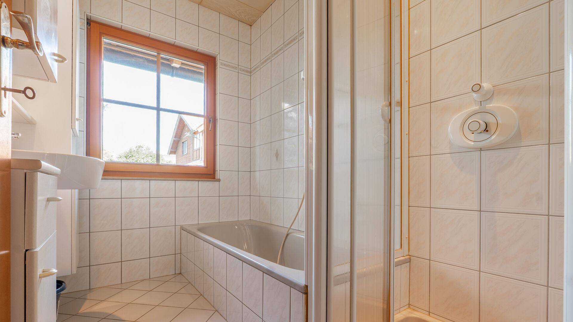 Badezimmer mit Fliesen, Badewanne mit Duschabtrennung, Waschbecken und Fenster.