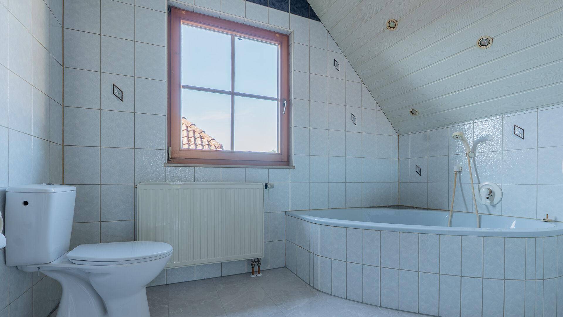 Badezimmer mit Fliesen, Eckbadewanne, Toilette und Fenster unter einer Dachschräge.