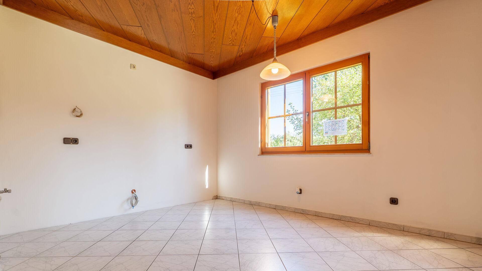 Leerer Raum, vorbereitet für eine Küche, mit Fliesenboden und Holzdecke, hell durch Fenster.