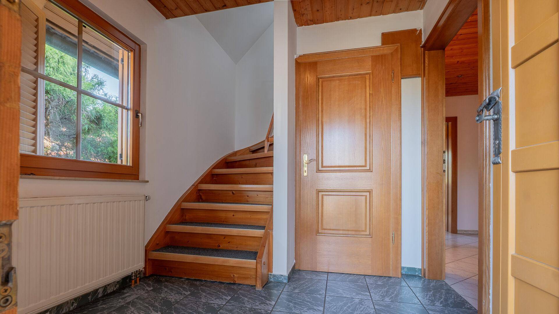 Heller Flur mit Fliesenboden, Holztreppe und Fenster, das Tageslicht hereinlässt.