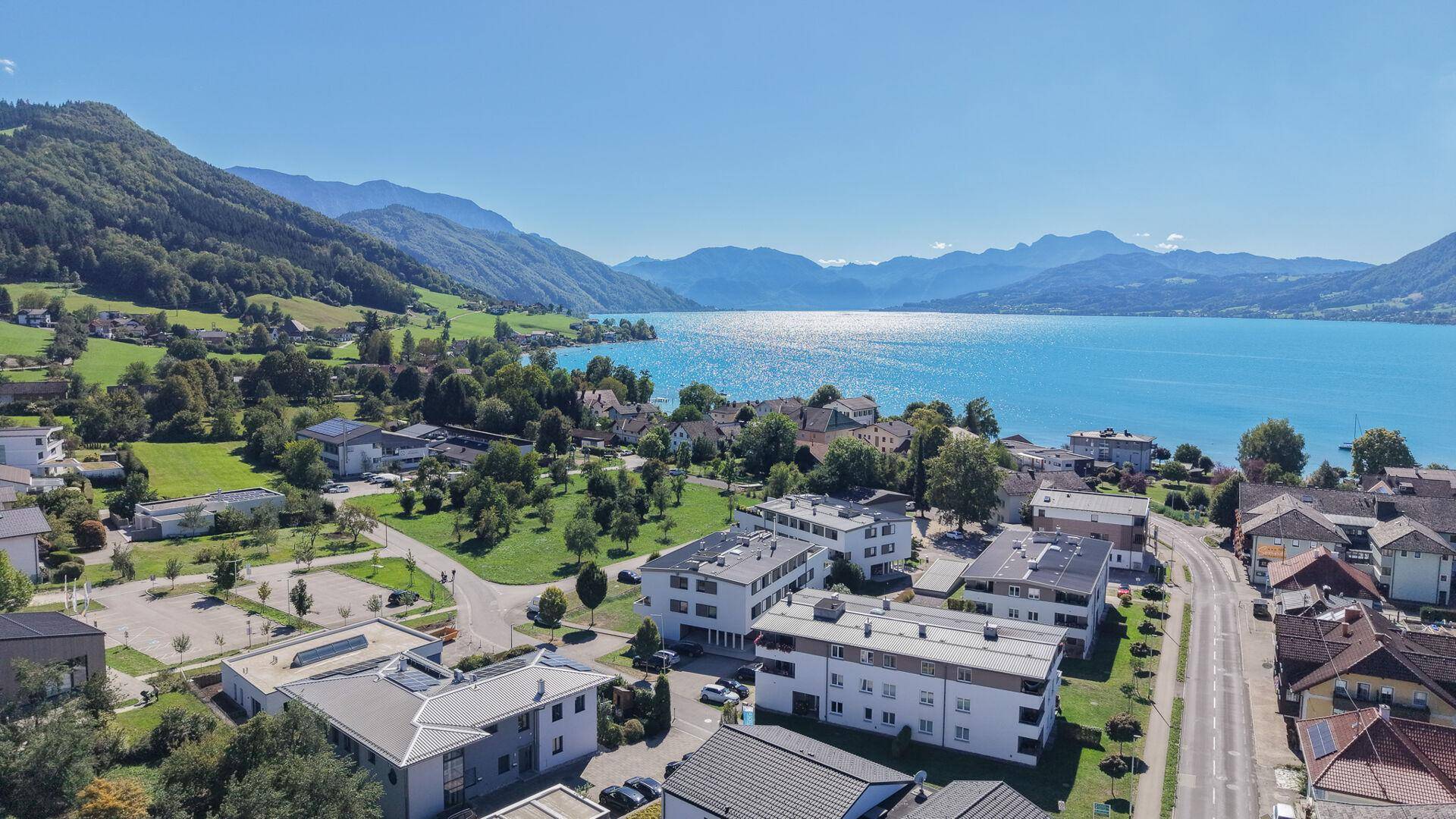 Weitläufige Luftaufnahme der Wohngegend mit See, Bergen und umliegender grüner Landschaft unter blauem Himmel.
