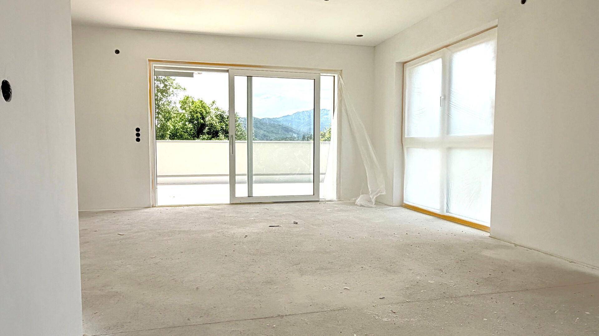 Geräumiger Wohnbereich im Rohbau mit großen Fenstern und Zugang zu einem Balkon mit Bergblick.