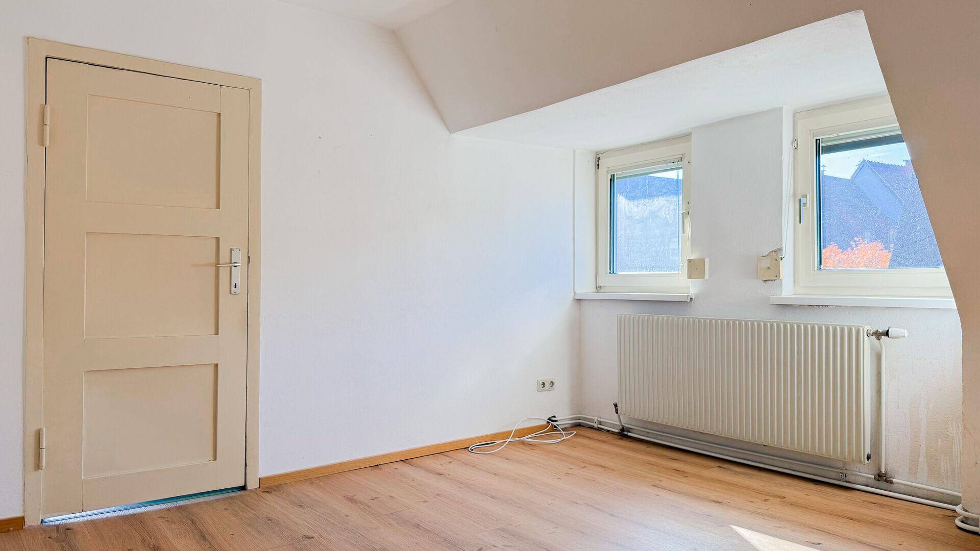 Leerer Dachbodenraum mit hellem Laminatboden, weißen Wänden und zwei Fenstern.
