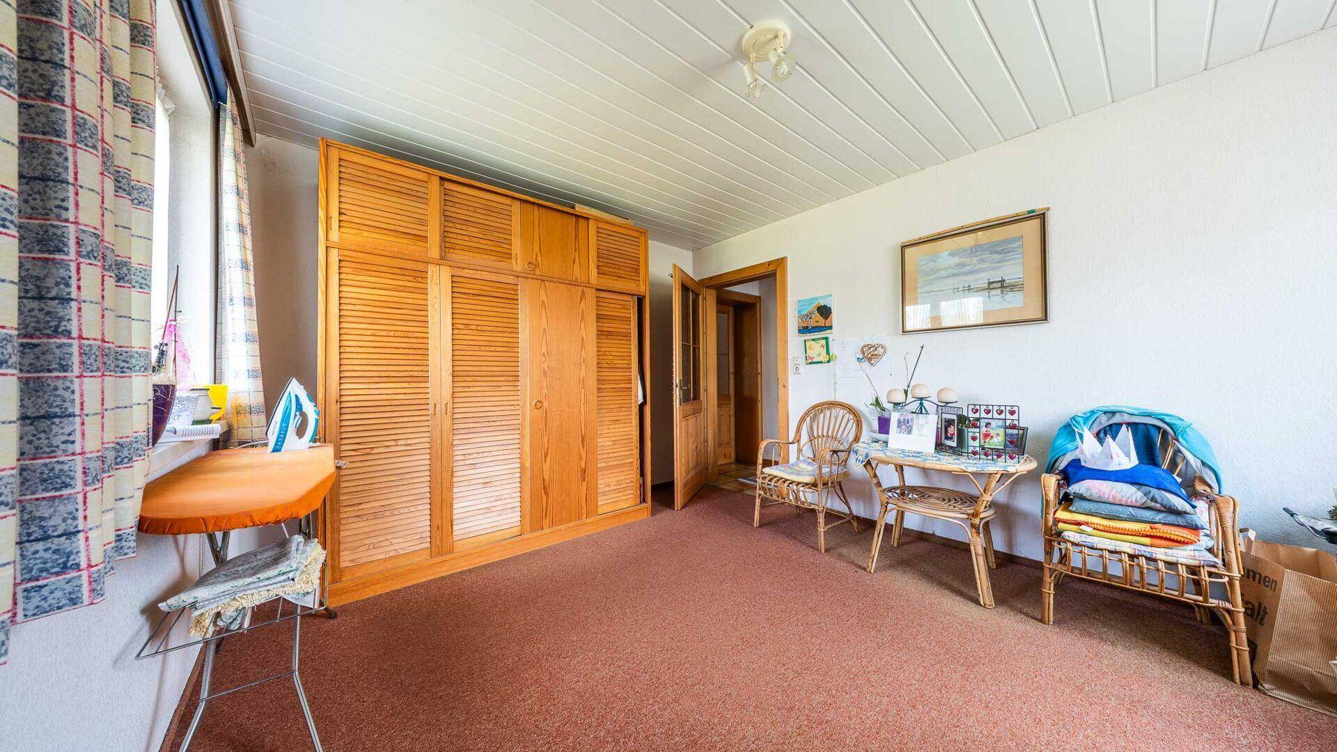 Gemütliches Schlafzimmer mit großem Holzschrank, Teppichboden und einem kleinen Sitzbereich am Fenster.