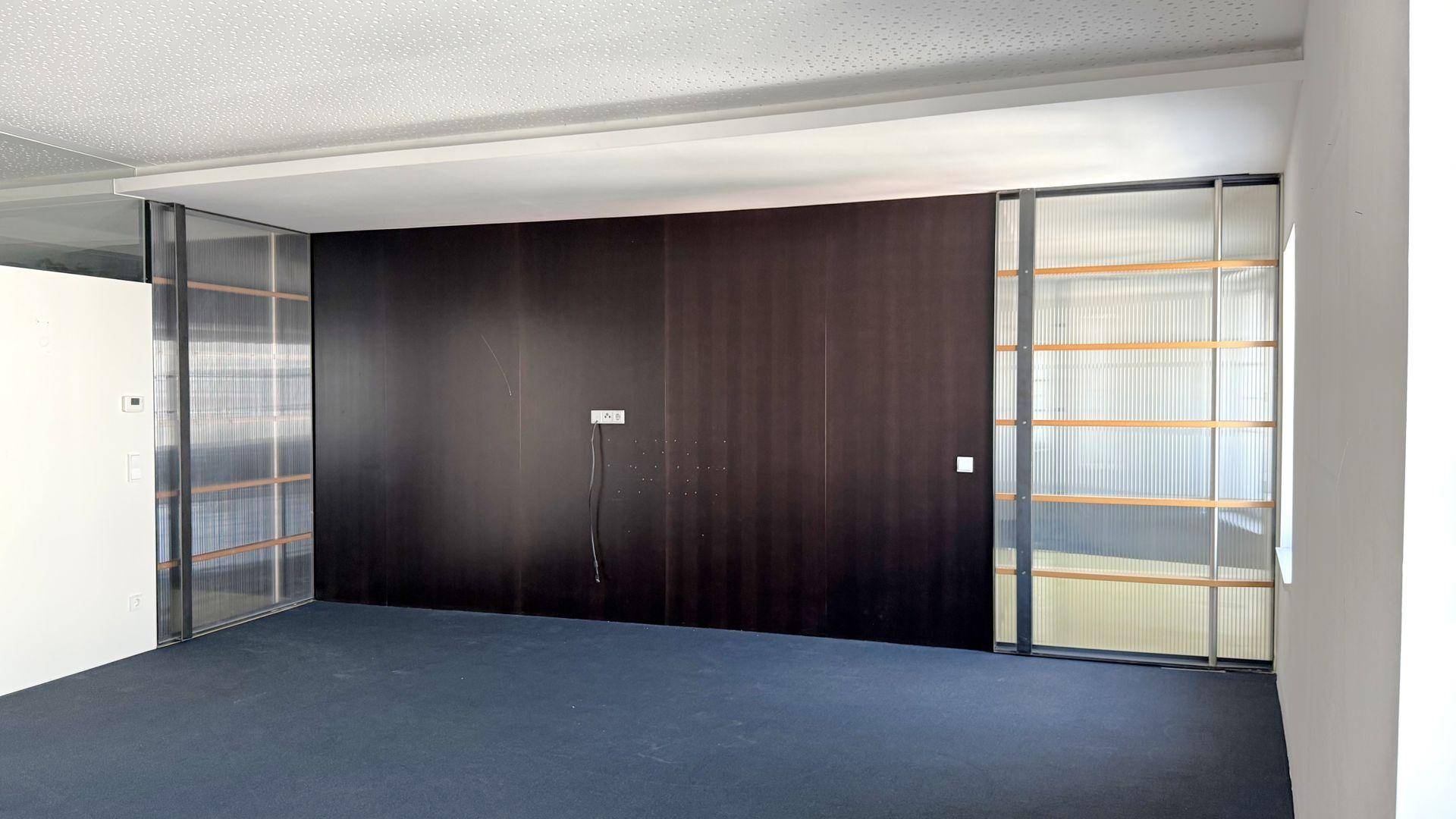 Großer, leerer Raum mit dunklem Teppichboden, einer dunklen Holzwand und eleganten Glasabtrennungen mit Holzrahmen.