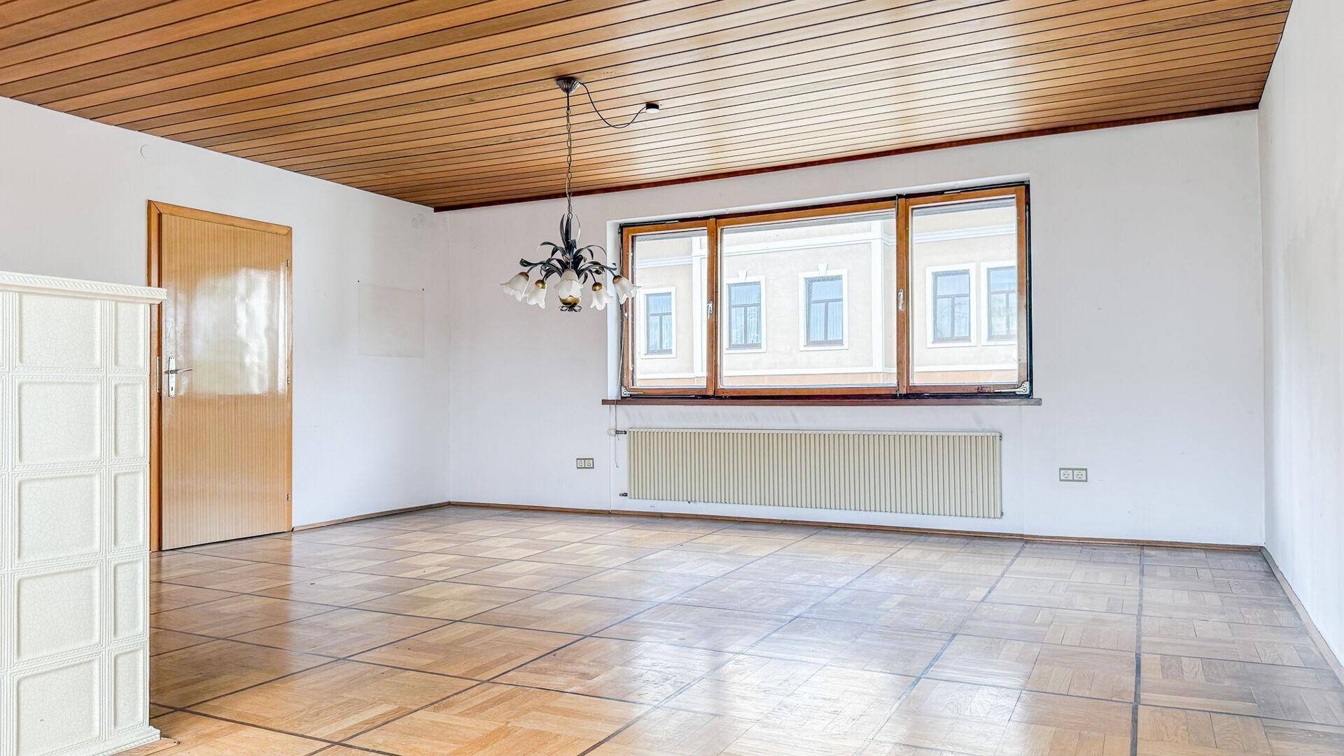 Leeres Wohnzimmer mit Fischgrätparkett, Holzdecke, großem Fenster und einem Kachelofen.