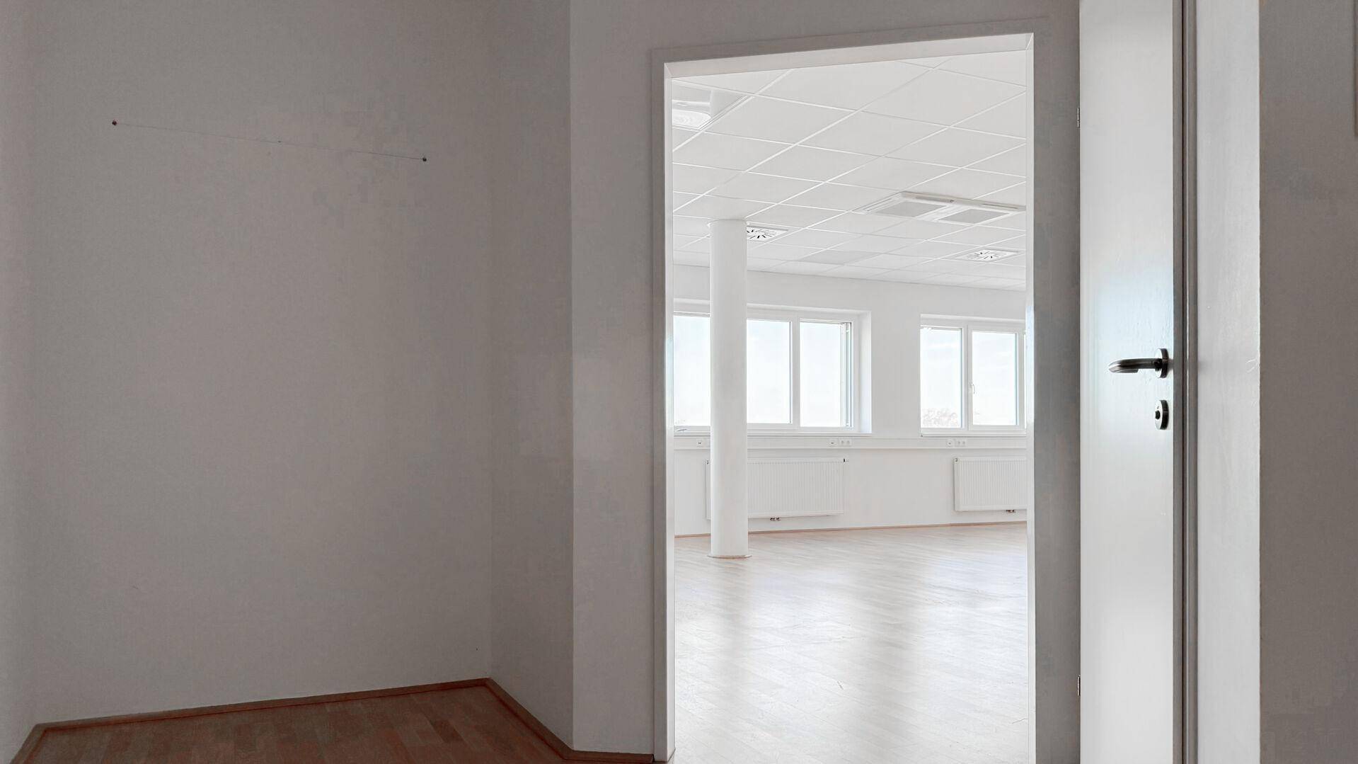 Blick aus einem Nebenraum in den hellen, leeren Bürobereich mit großen Fenstern.