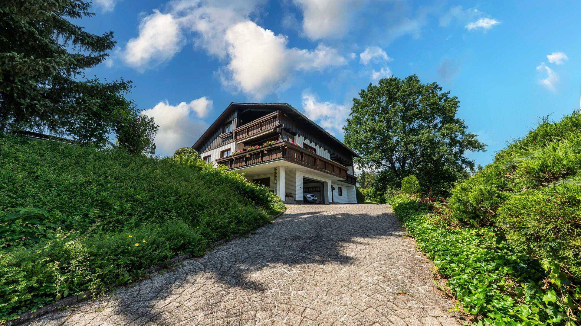 Das Haus von der gepflasterten Auffahrt aus gesehen, umgeben von viel Grün und Bäumen.