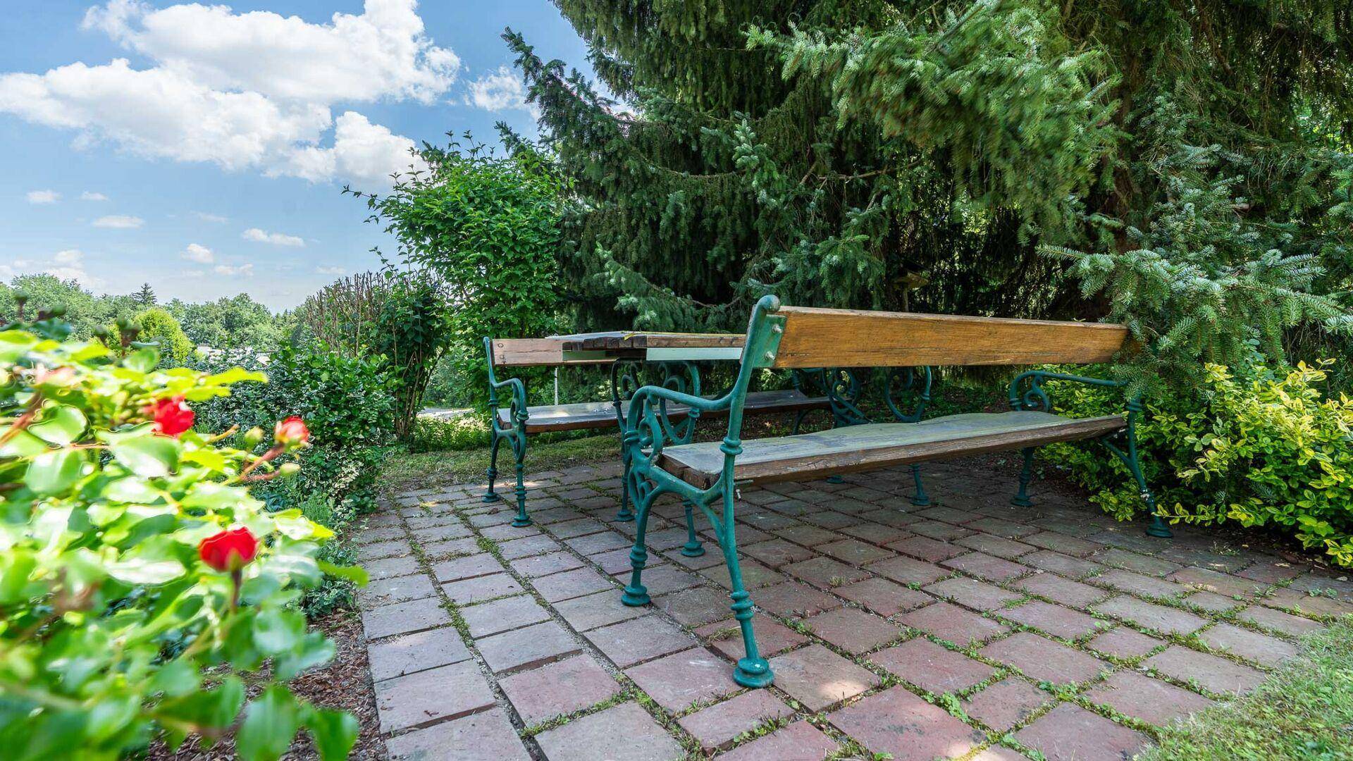 Gemütlicher Sitzbereich im Garten mit rustikalen Bänken und einem gepflasterten Boden, umgeben von Blumen.