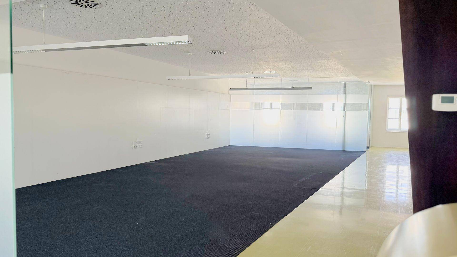 Geräumiger, leerer Büroraum mit dunklem Teppichboden, weißen Wänden und modernen Glasabtrennungen für flexible Nutzung.