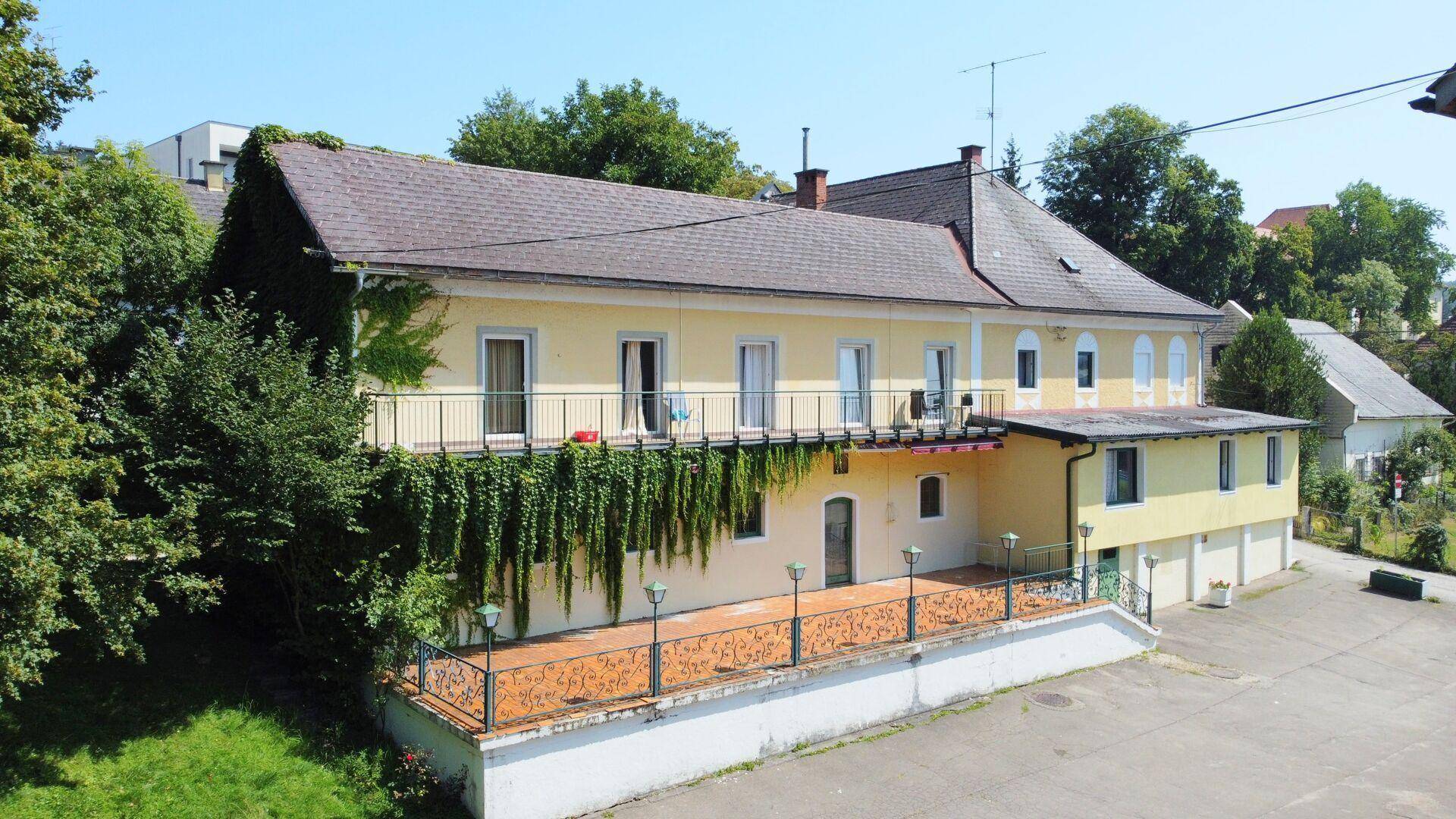 Seitenansicht des Gebäudes mit einer großen Terrasse und einem Balkon, umgeben von üppigem Grün.