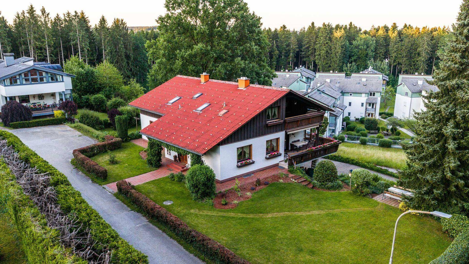 Luftaufnahme des Hauses mit rotem Dach, umgeben von Wald und weiteren Wohnhäusern.