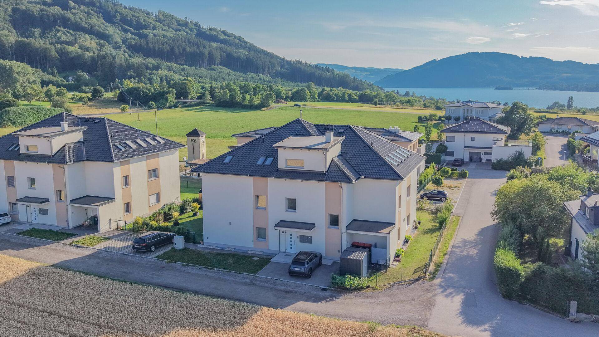 Luftaufnahme einer modernen Wohnanlage mit mehreren Häusern und Seeblick unter blauem Himmel.