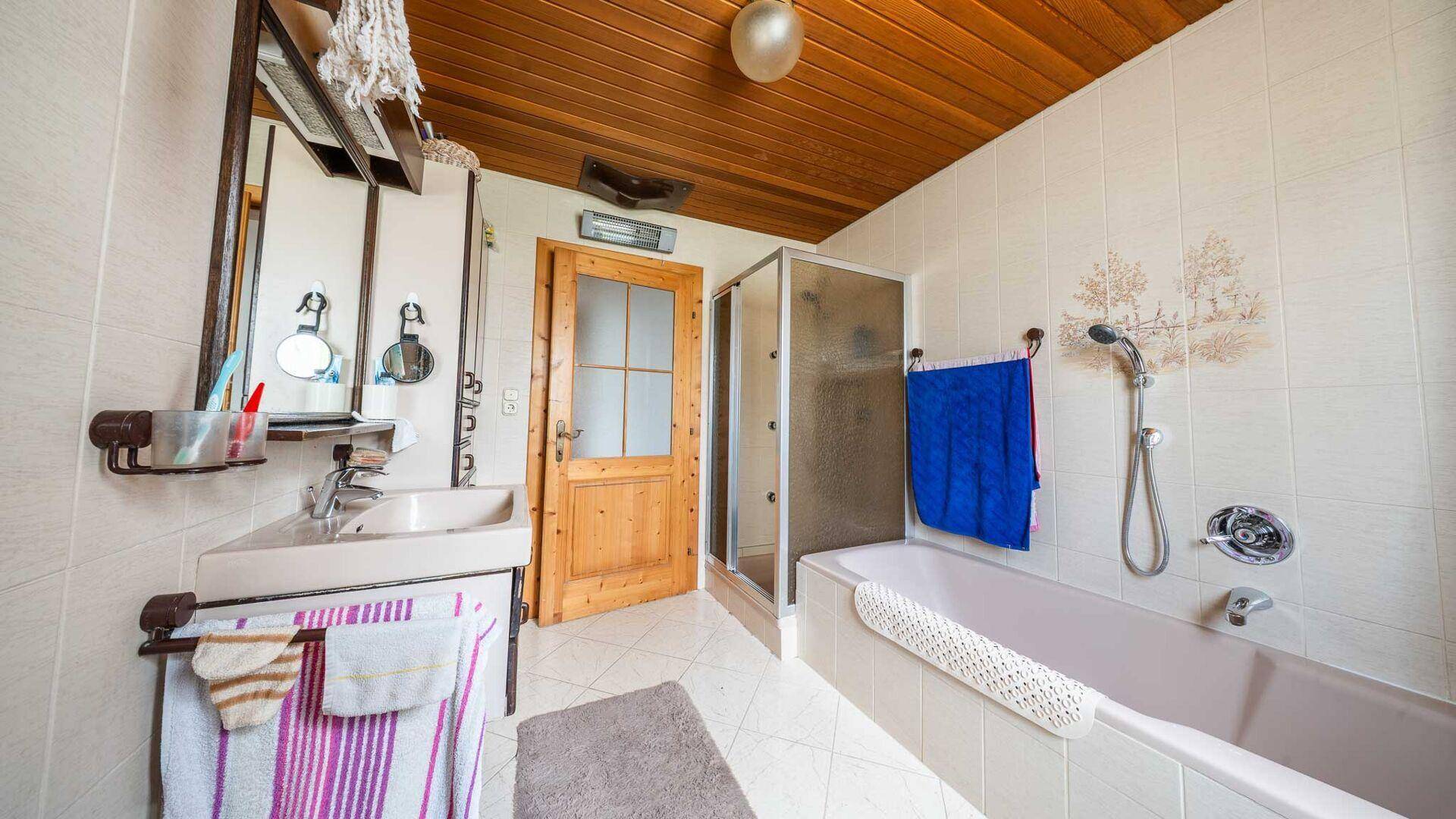 Funktionales Badezimmer mit Badewanne, Duschkabine und Holzdecke, ausgestattet mit einfachen Fliesen.