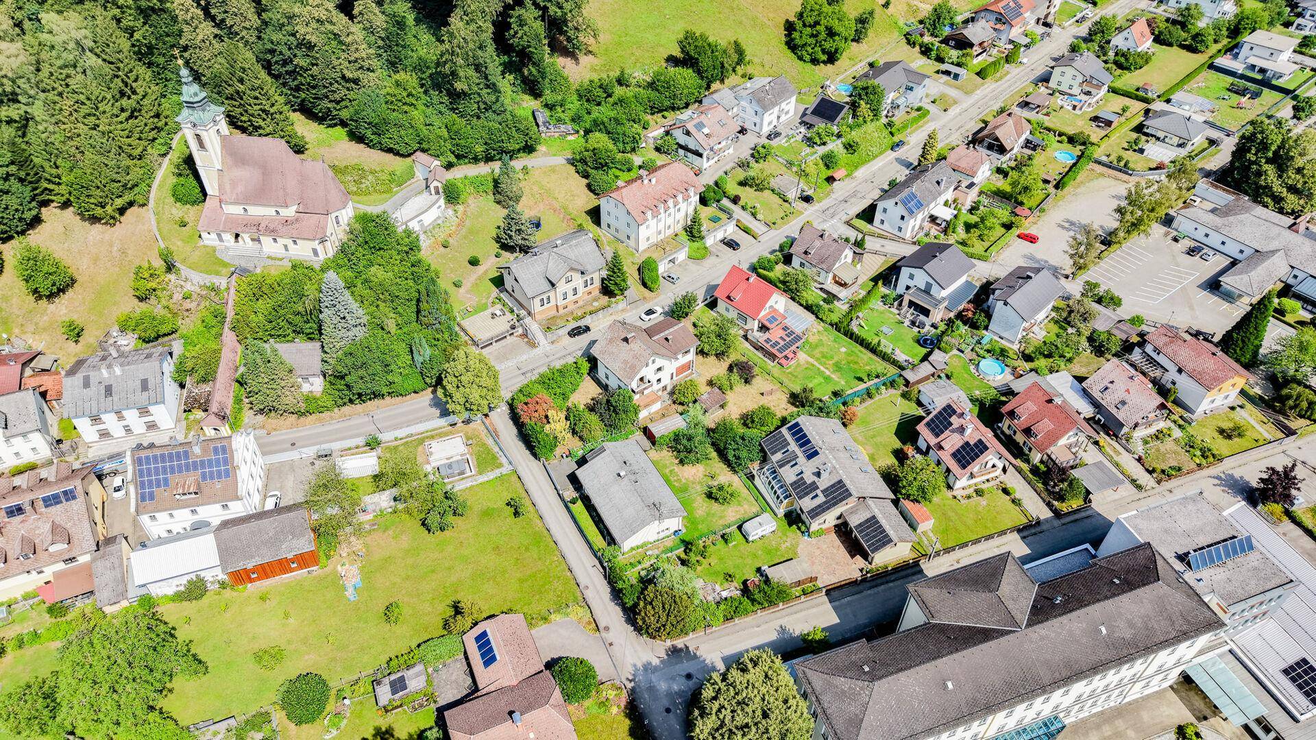 Panoramische Luftaufnahme des Dorfes mit Wohngebäuden, viel Grün und einer prominenten Kirche.