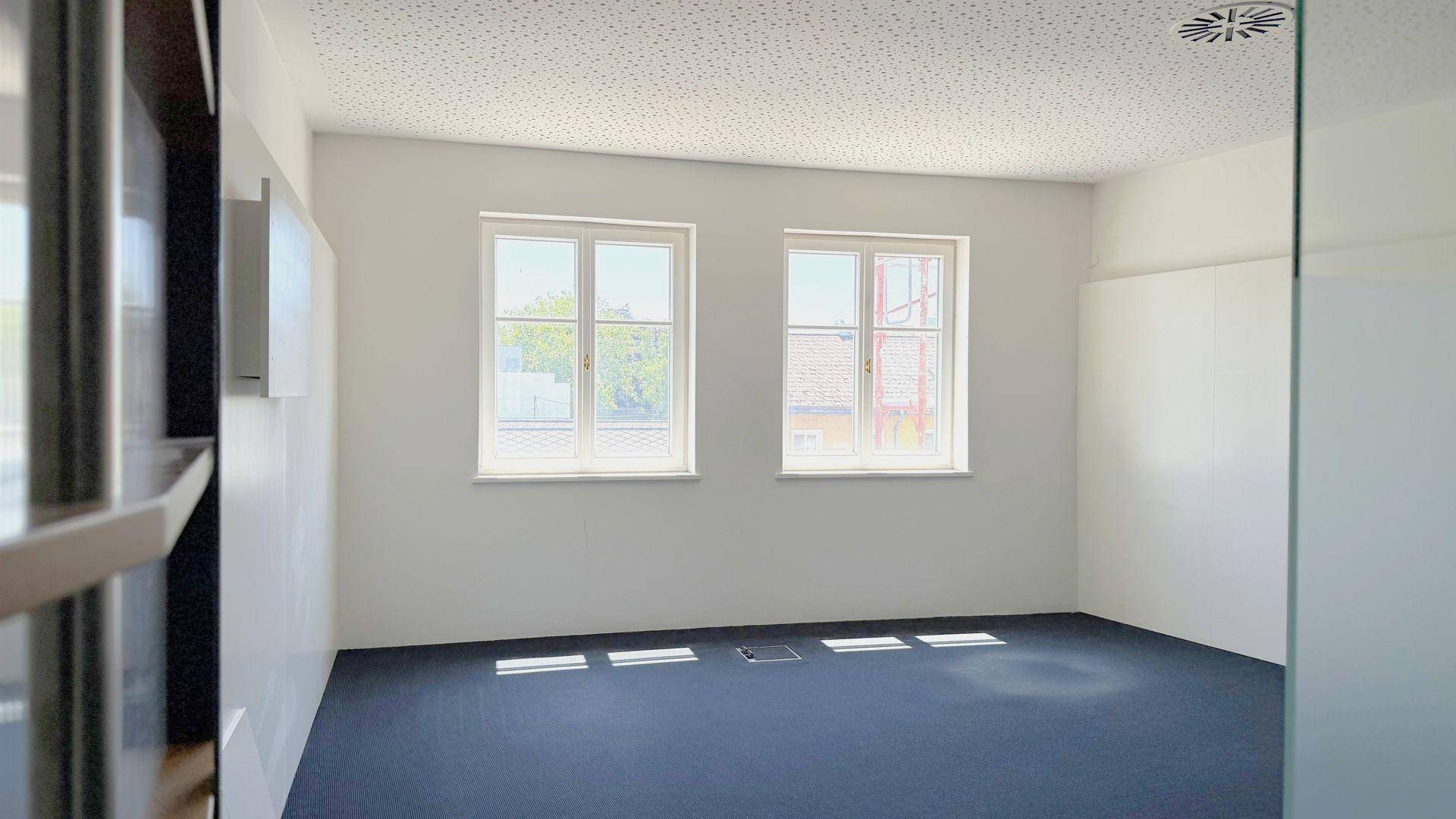 Helles, leeres Büro mit dunklem Teppichboden, weißen Wänden und zwei großen Fenstern, die viel Tageslicht hereinlassen.