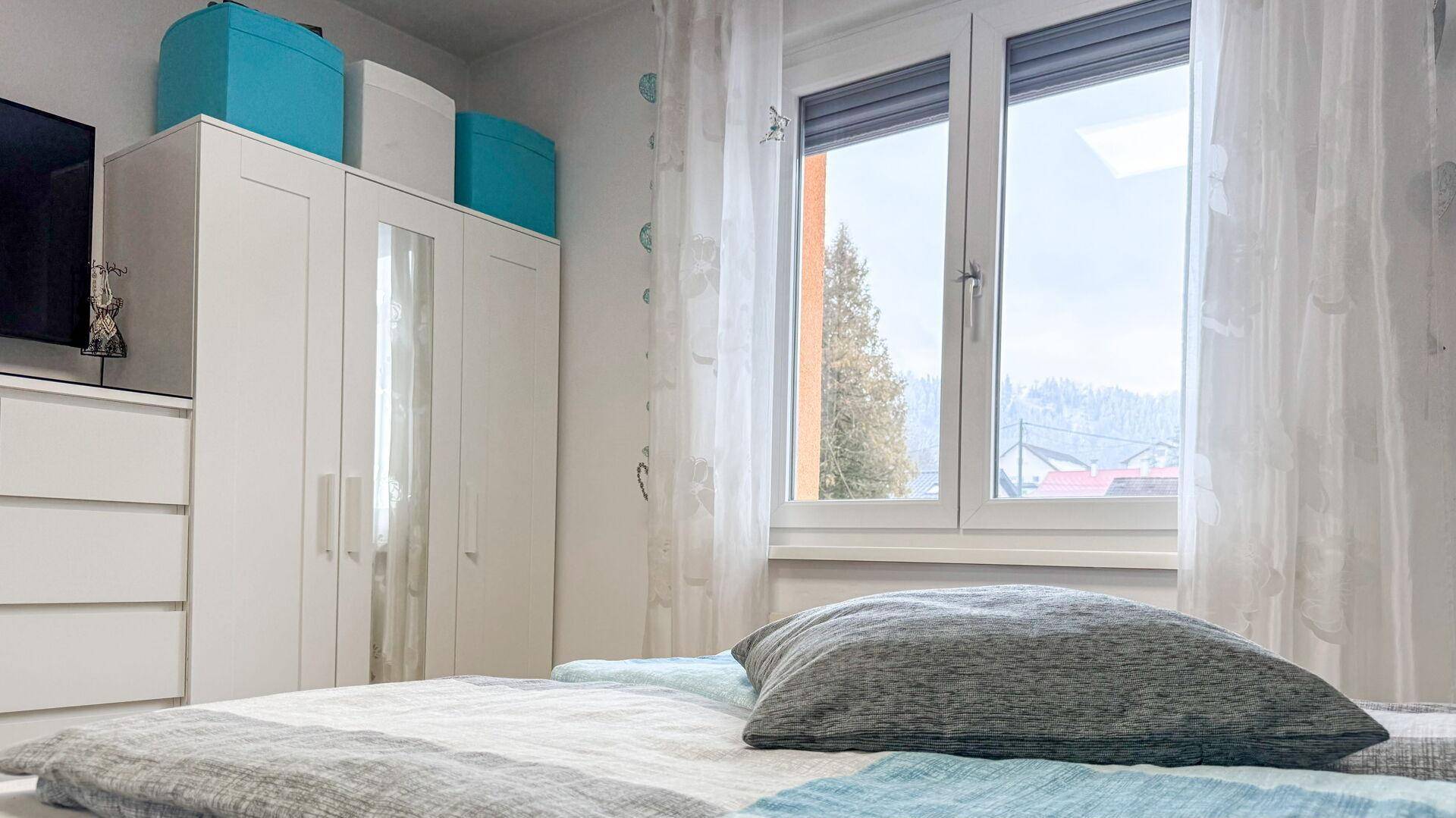 Schlafzimmer mit großem Fenster, Rollläden und Blick auf die umliegende Landschaft.