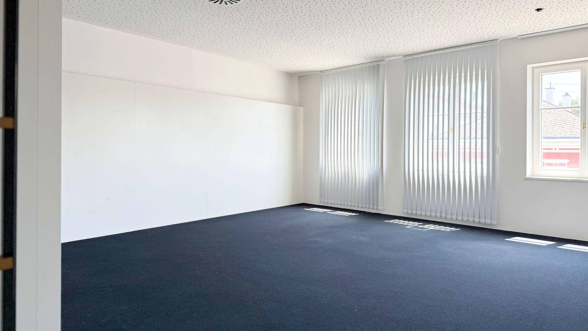 Geräumiges, leeres Büro mit dunklem Teppichboden, weißen Wänden und Fenstern, die mit vertikalen Jalousien ausgestattet sind.