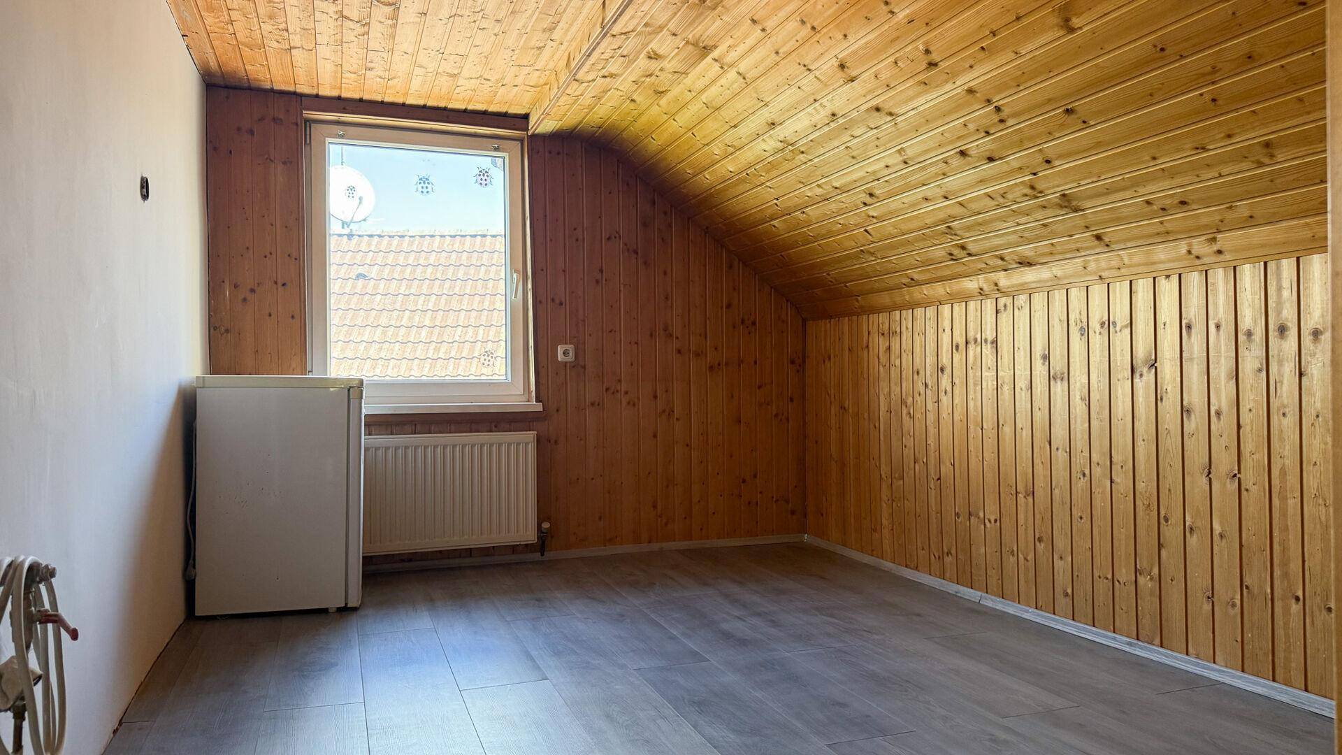 Dachzimmer mit Holzverkleidung, schräger Decke, Fenster und einem kleinen Kühlschrank.