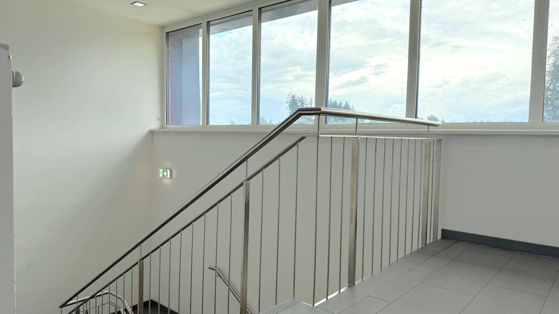 Modernes Treppenhaus mit Edelstahlgeländer, großen Fenstern und hellen Fliesen, die viel Licht hereinlassen.