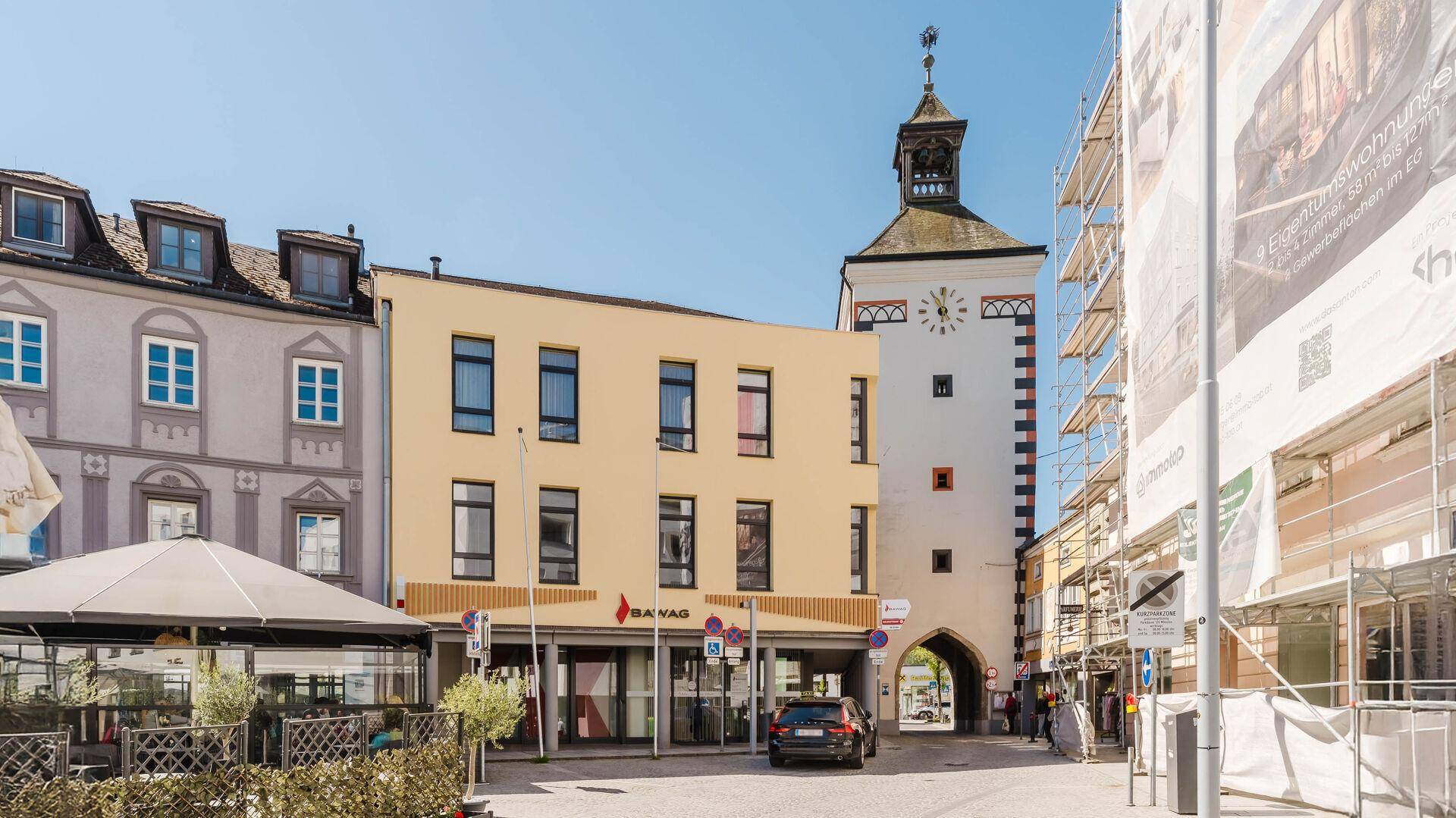 Historisches Gebäude mit Uhrturm und angrenzendem modernen Bau in einer belebten Straße.