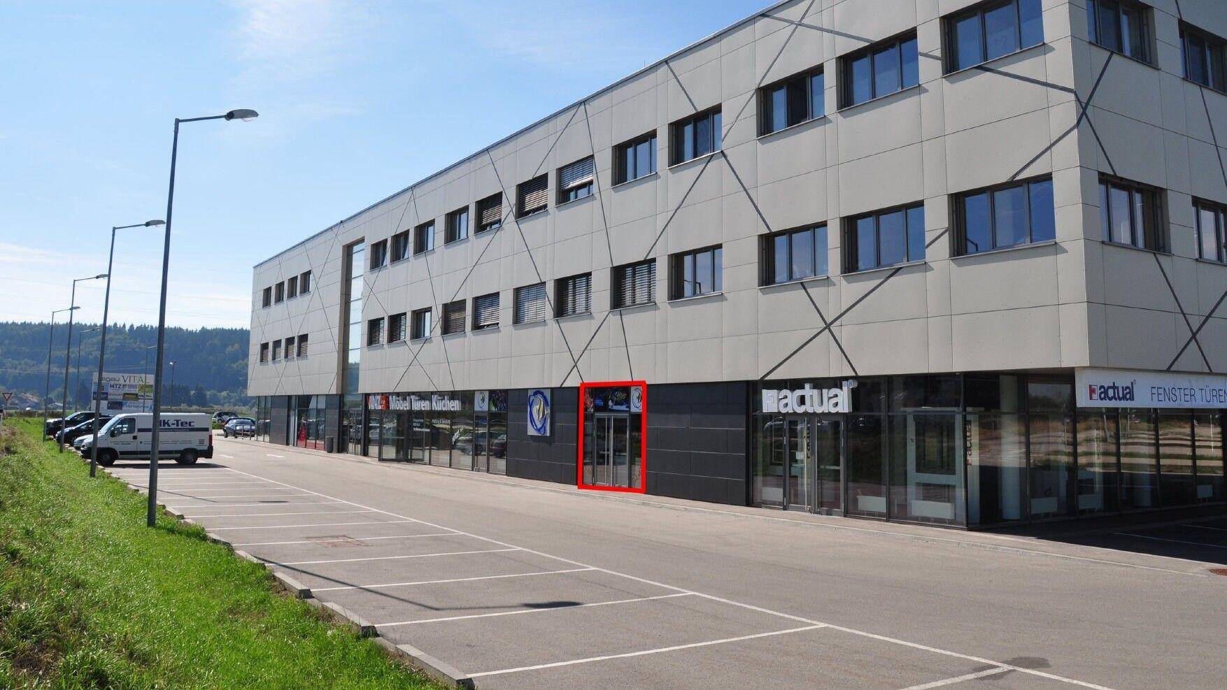 Moderne Geschäftsfassade mit großen Fenstern und einem markierten Eingangsbereich an einem sonnigen Tag.