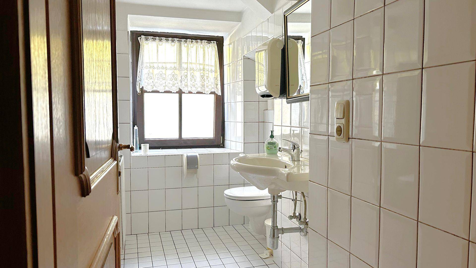 Toilettenraum mit Fenster, Waschbecken und weißen Fliesen an Wänden und Boden.