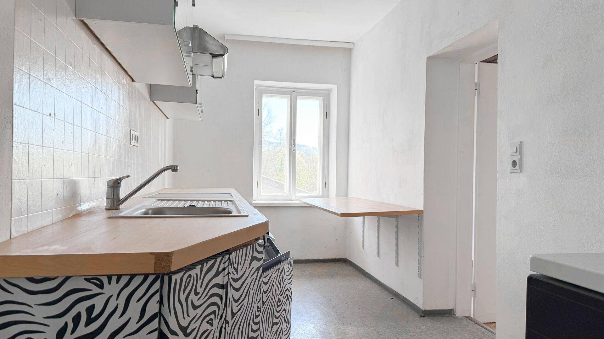 Einfache Küche mit Zebra-Muster-Fronten, Spüle und einem kleinen Klapptisch am Fenster.