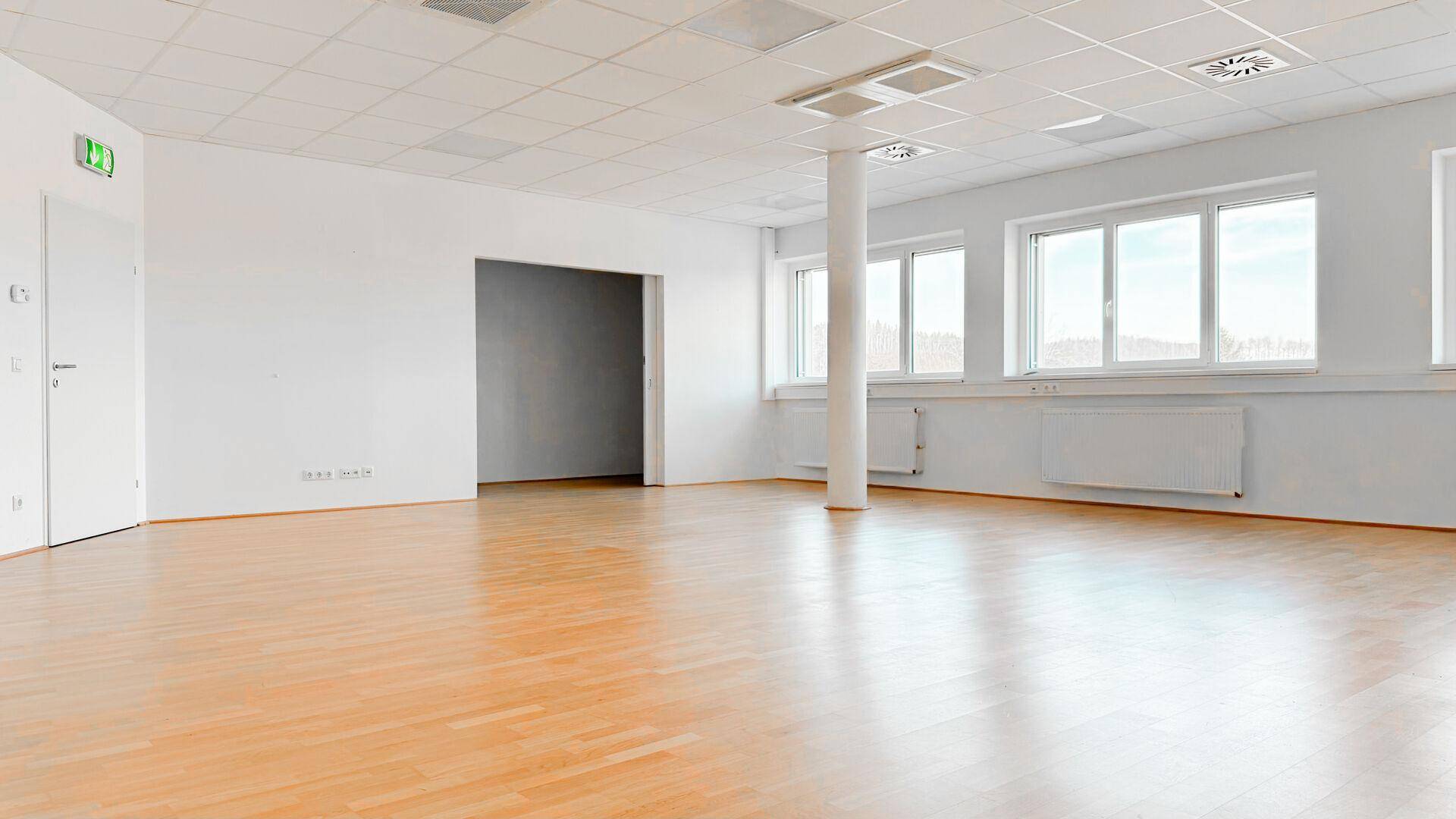 Leerer Büroraum mit Holzboden, großen Fenstern und Zugang zu einem weiteren Bereich.