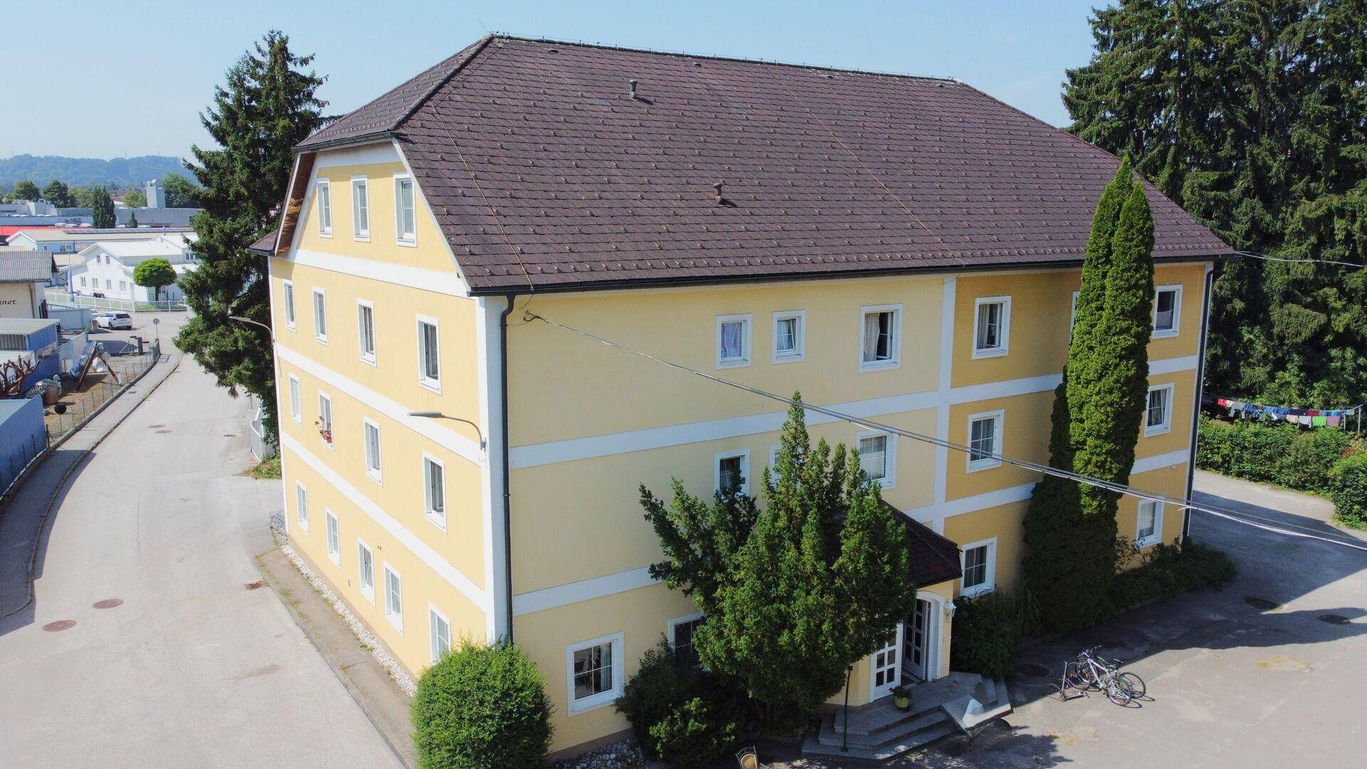 Ansicht des mehrstöckigen Gebäudes mit gelber Fassade, weißen Fenstern und hohen Bäumen.