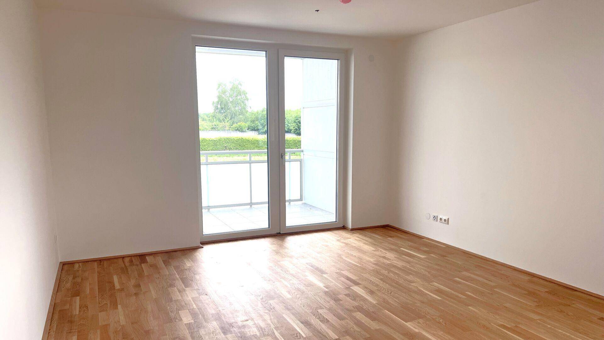 Helles Zimmer mit Holzboden und Zugang zu einem Balkon, umgeben von grüner Landschaft.