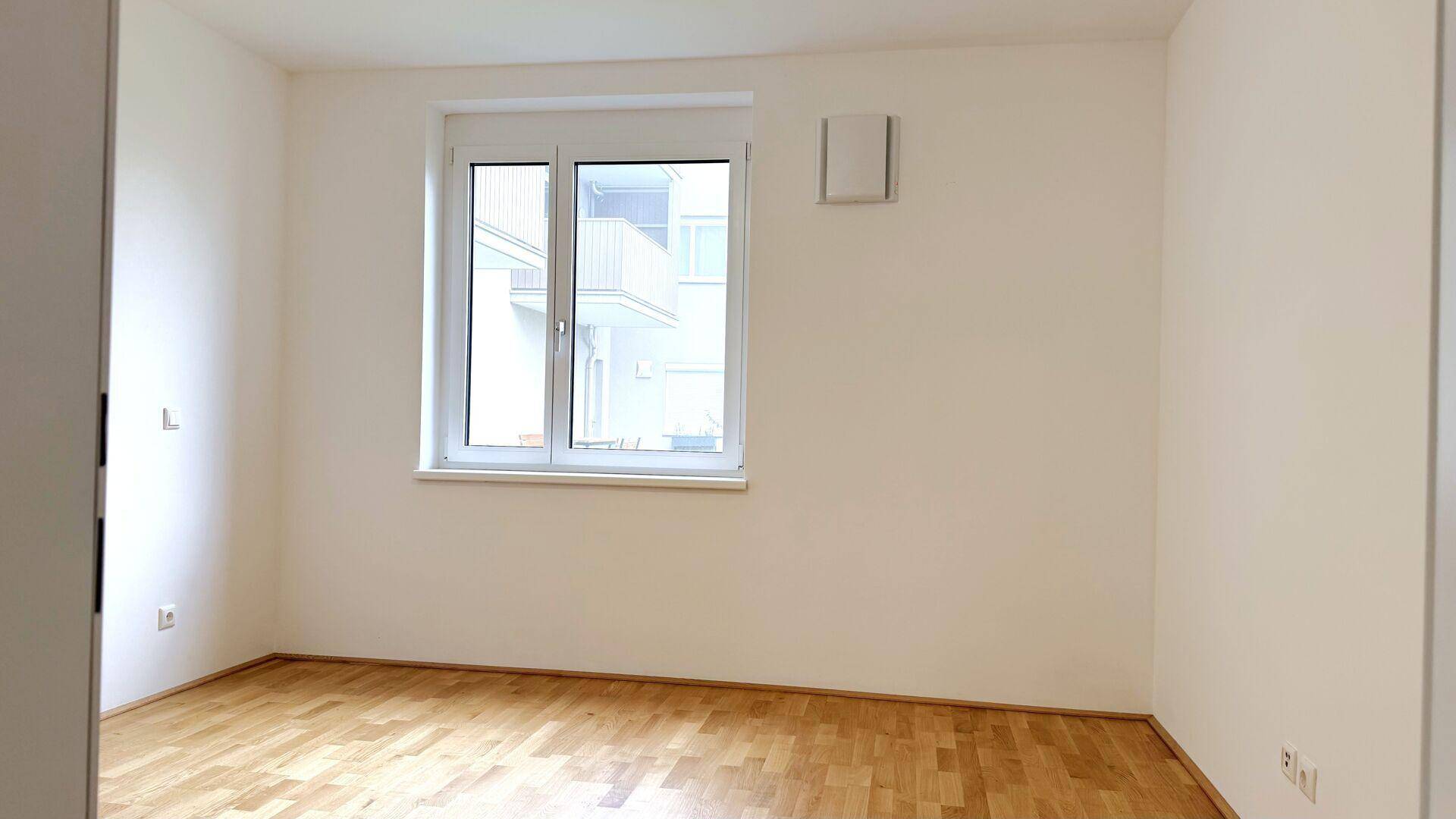 Freundliches Zimmer mit Parkettboden und einem großen Fenster, das den Blick auf den Innenhof freigibt.