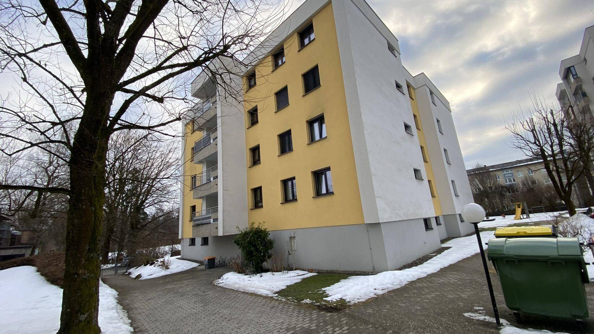 Mehrstöckiges Wohngebäude mit gelber und weißer Fassade, umgeben von Bäumen und Schnee.