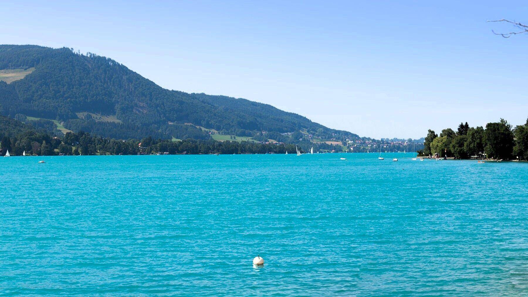 Weitläufiger Blick über einen türkisblauen See mit bewaldeten Bergen und Uferbebauung in der Ferne.